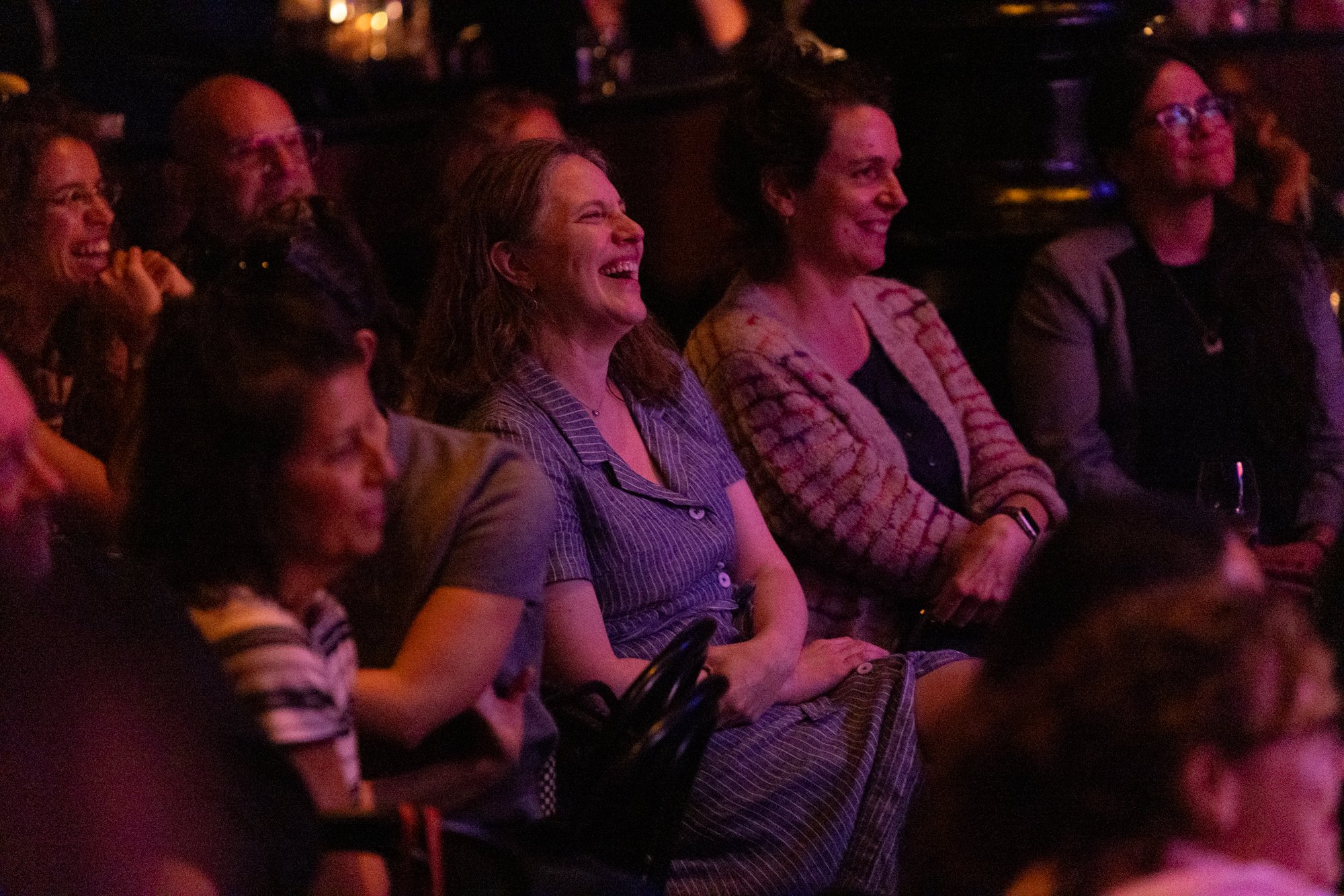 An audience of adults watching a performance or presentation, laughing and smiling in a dimly lit venue.