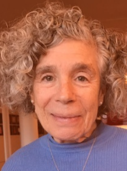 Elderly woman with curly hair smiling indoors wearing a blue shirt.