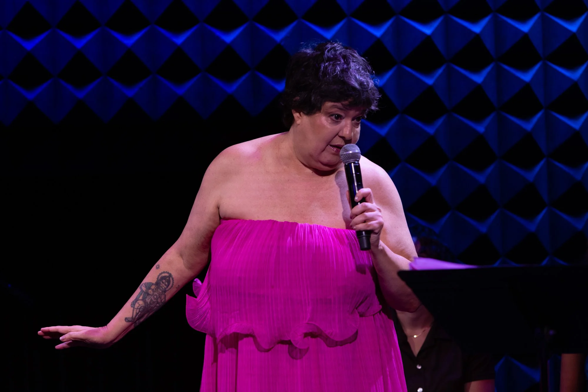 A woman with short, curly hair and tattoos on her arms is wearing a bright pink strapless dress and holding a microphone, standing on stage with a dark background that has a blue, geometric, pillow-like pattern.