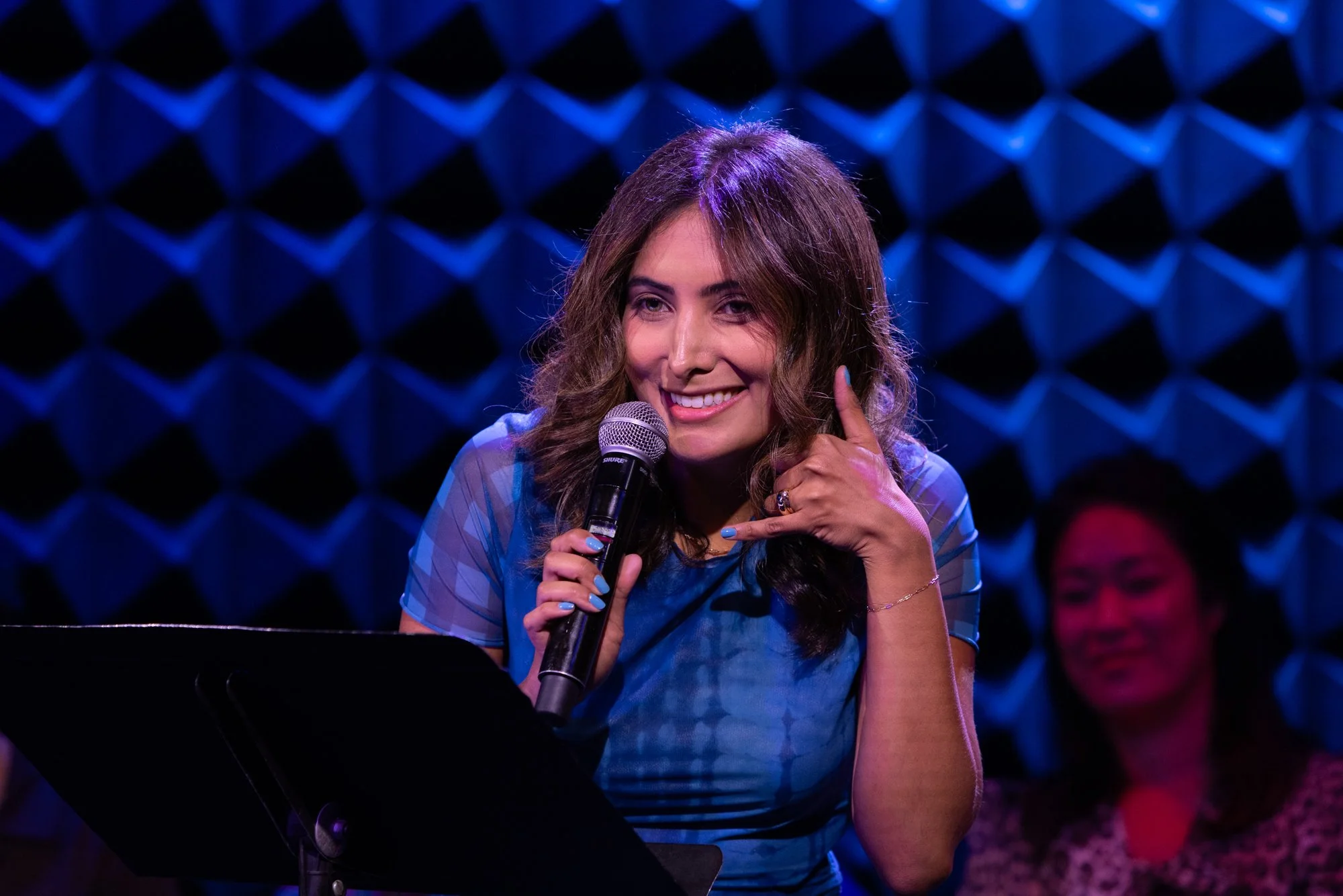 A woman with long brown hair, wearing a blue top, smiling, holding a microphone, in a soundproofed studio with blue acoustic panels in the background.