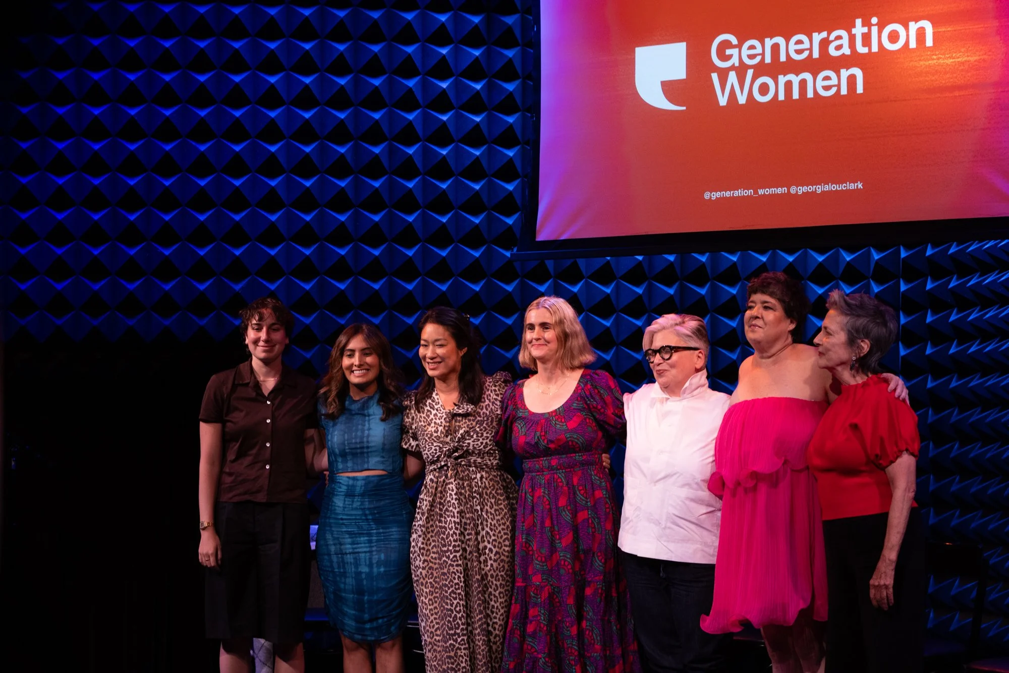 A group of women standing together on stage at a Generation Women event, with a large screen behind them displaying the event's logo and social media handles.