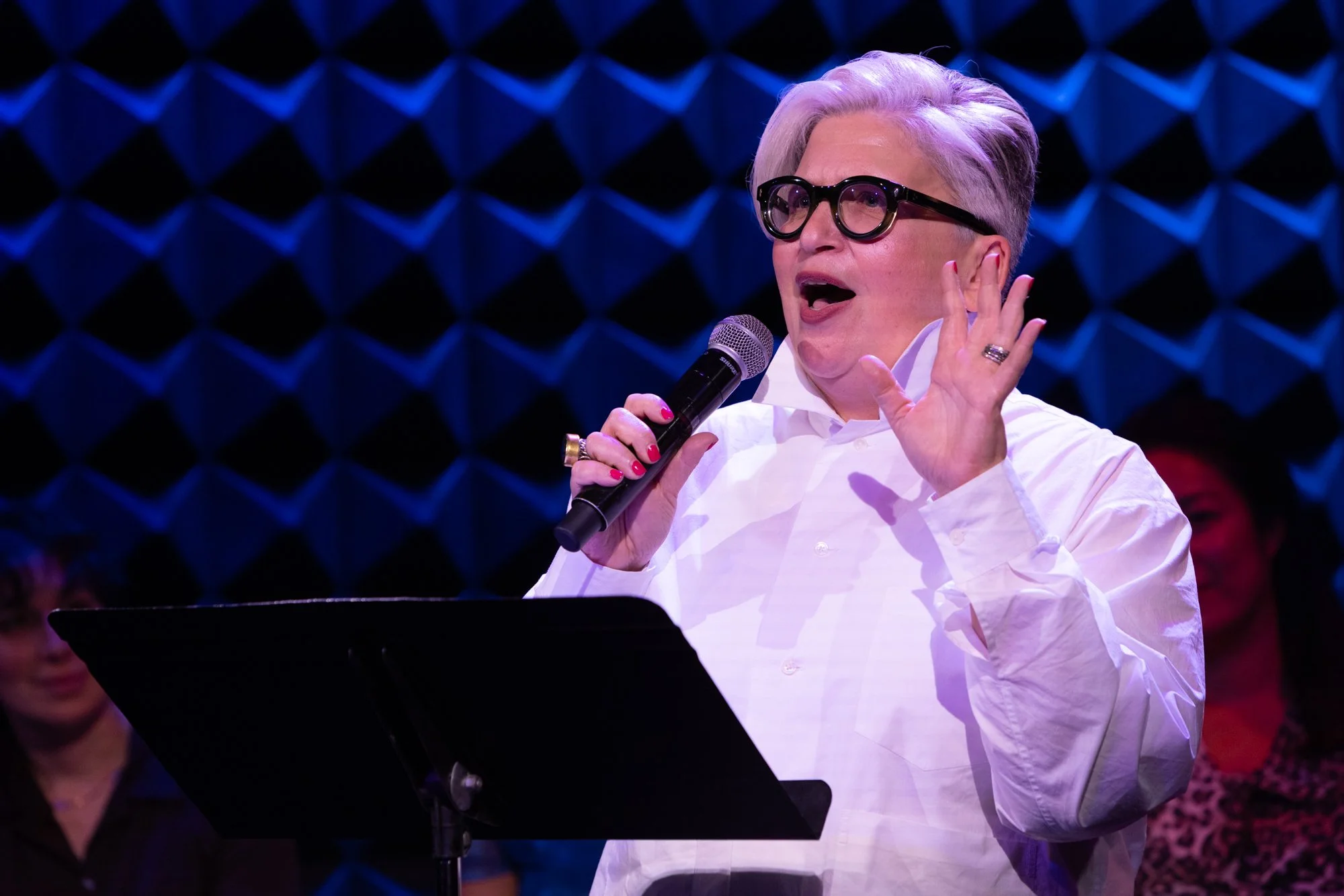 A woman with short gray hair and black glasses singing into a microphone, wearing a white shirt, with a blue and black geometric background and a music stand in front of her.