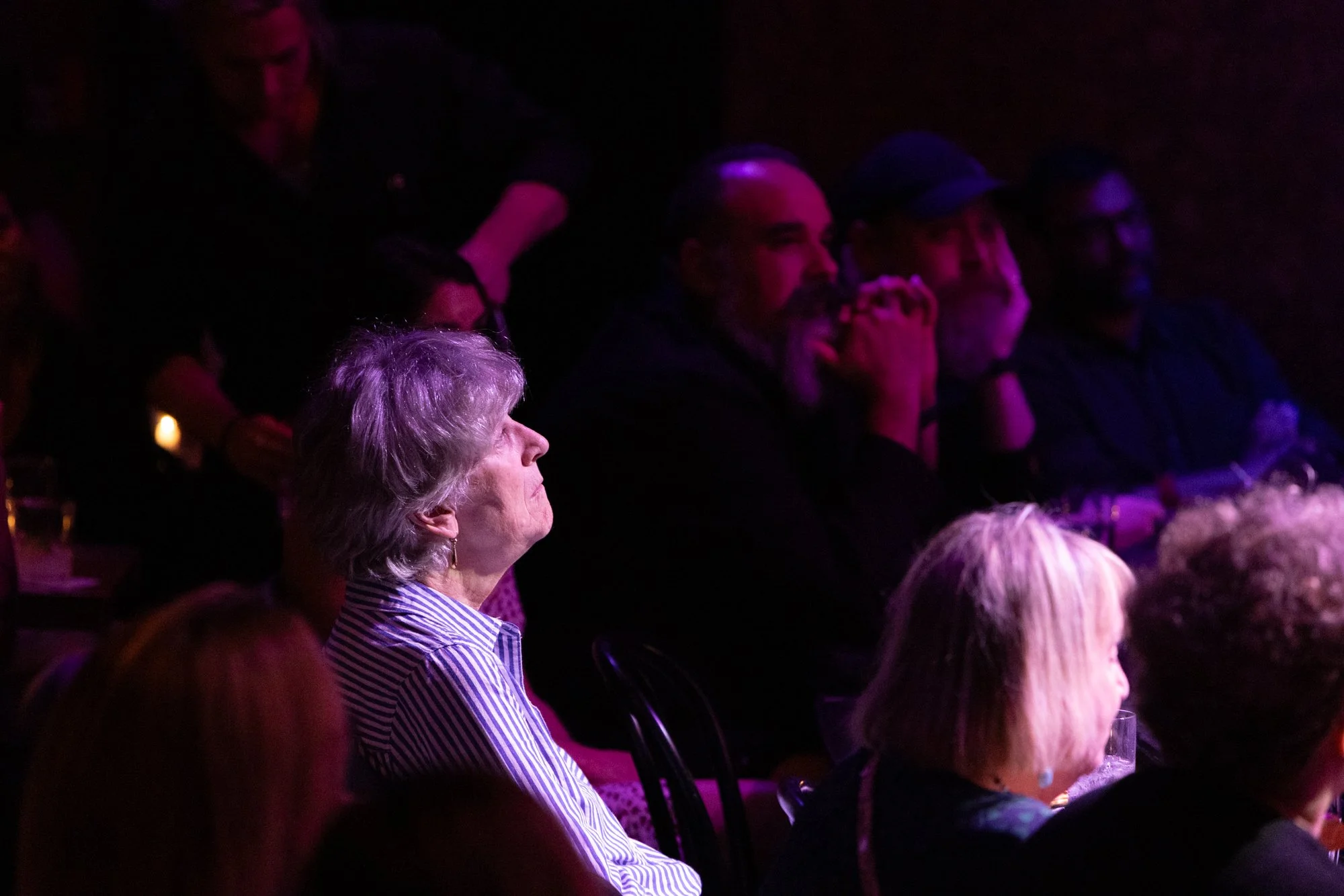 Audience attentively watching a performance or presentation in a darkened setting illuminated with purple and pink lighting.