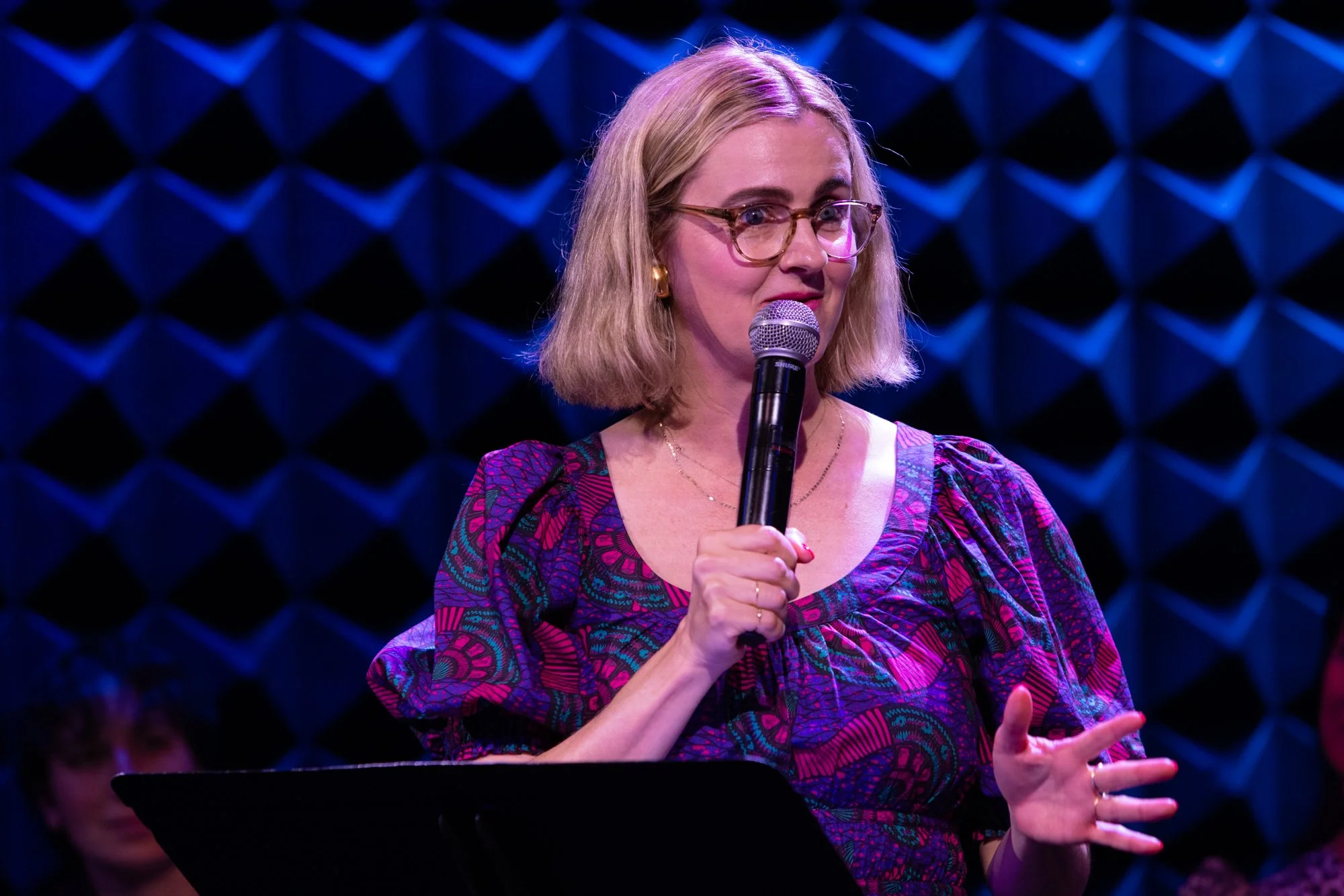 A woman with blonde hair, glasses, and hoop earrings is speaking into a microphone against a blue, geometric background, wearing a colorful patterned top.