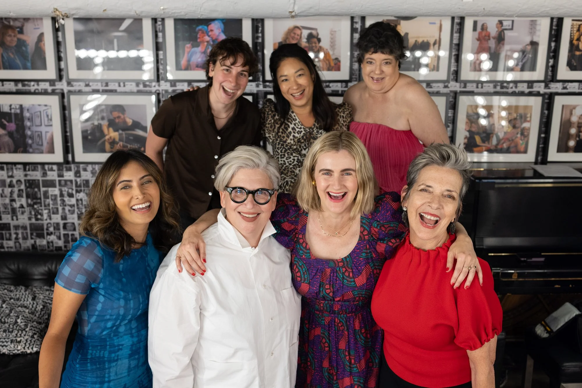 Group of nine smiling women and men posing together in a room with framed photographs on the wall.