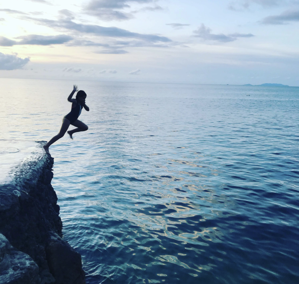 Person jumping off a rocky ledge into the water during sunset or early evening.