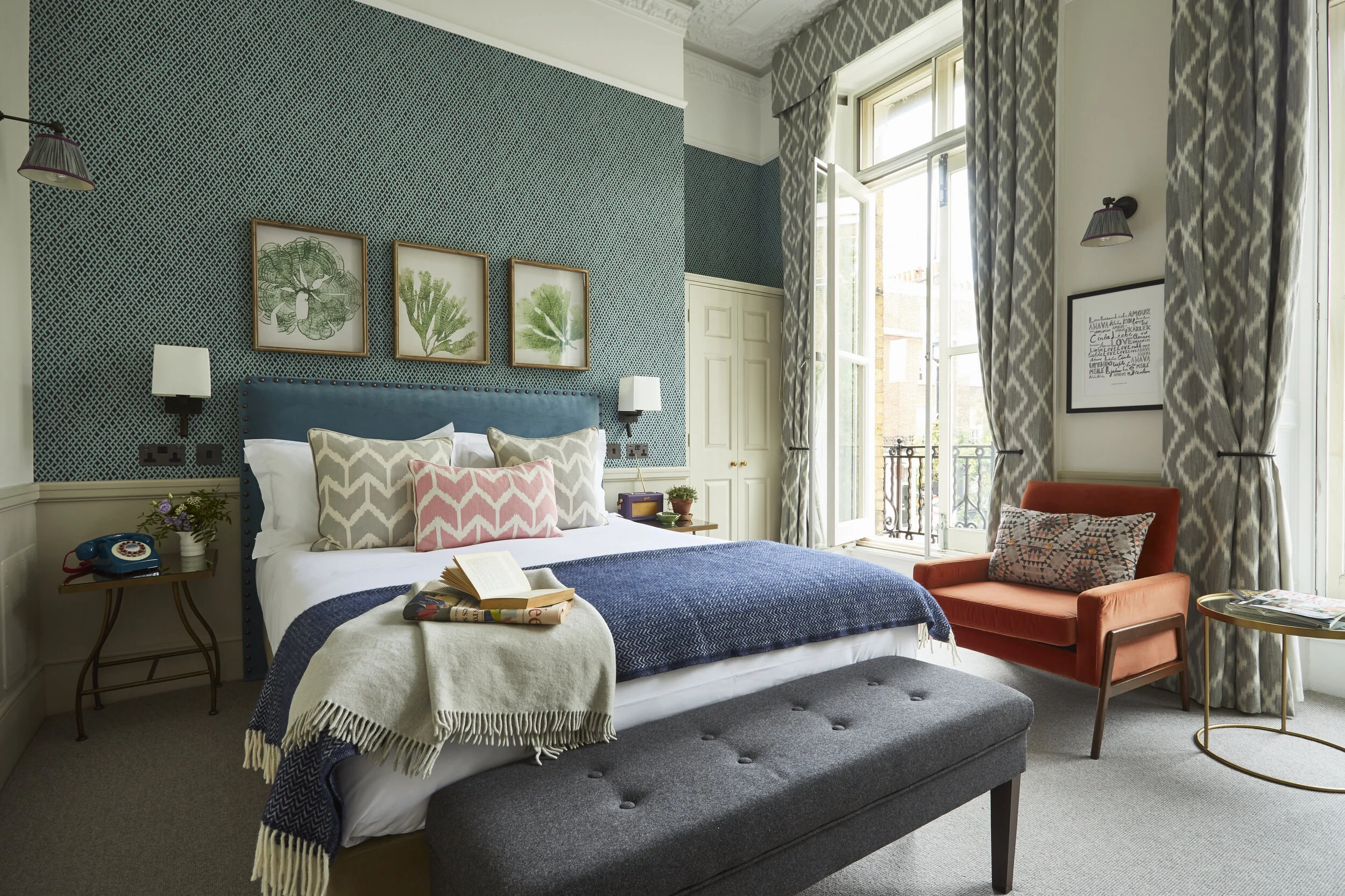 A cozy bedroom with a double bed featuring a blue headboard, decorative pillows, and a blanket. There are framed botanical prints above the bed, a bedside table with a vintage rotary phone, and a small sitting area with an orange armchair by tall win