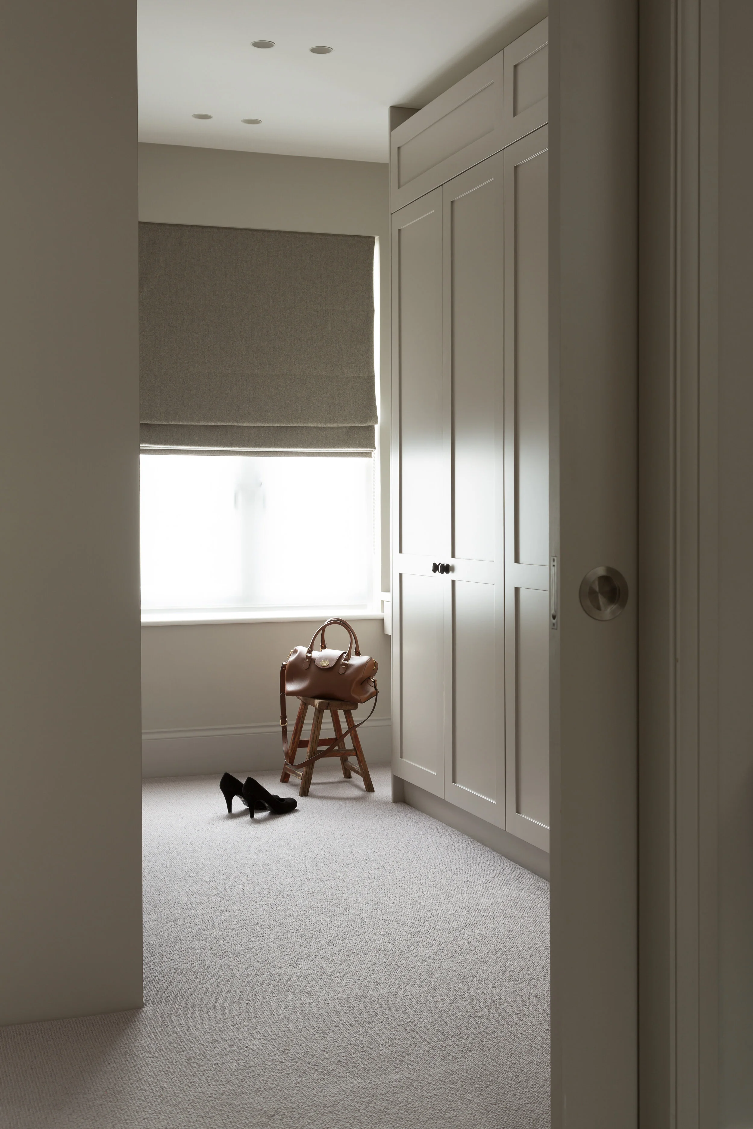 An interior view of a room with a window, beige carpet, white cabinetry, a brown handbag on a small wooden stool, and a pair of black high heels on the floor.