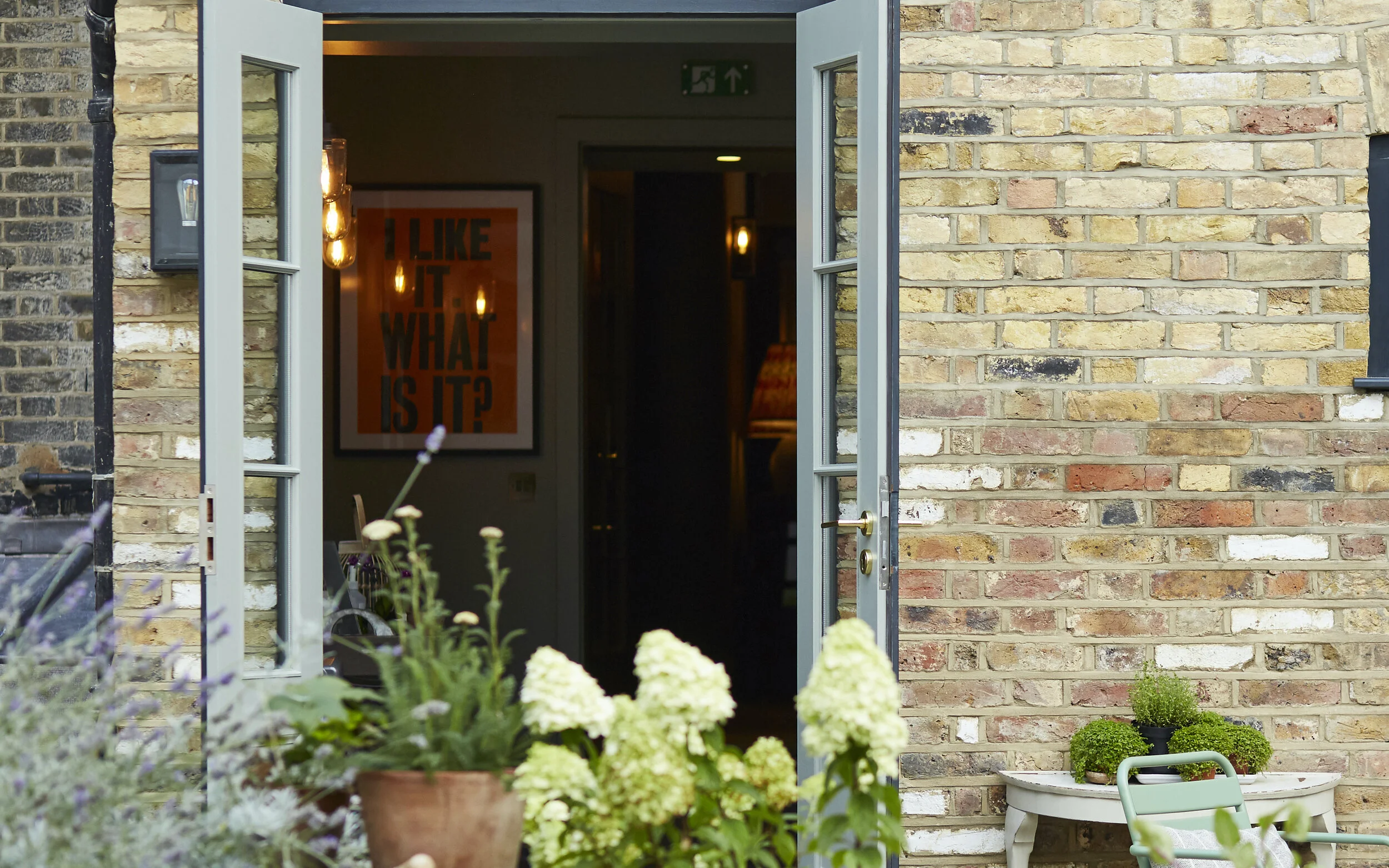 Open French door leading into a house with a brick exterior, featuring flowers and potted plants outside, and a prominent orange poster inside that reads 'I LIKE IT. WHAT IS IT?' with warm lighting