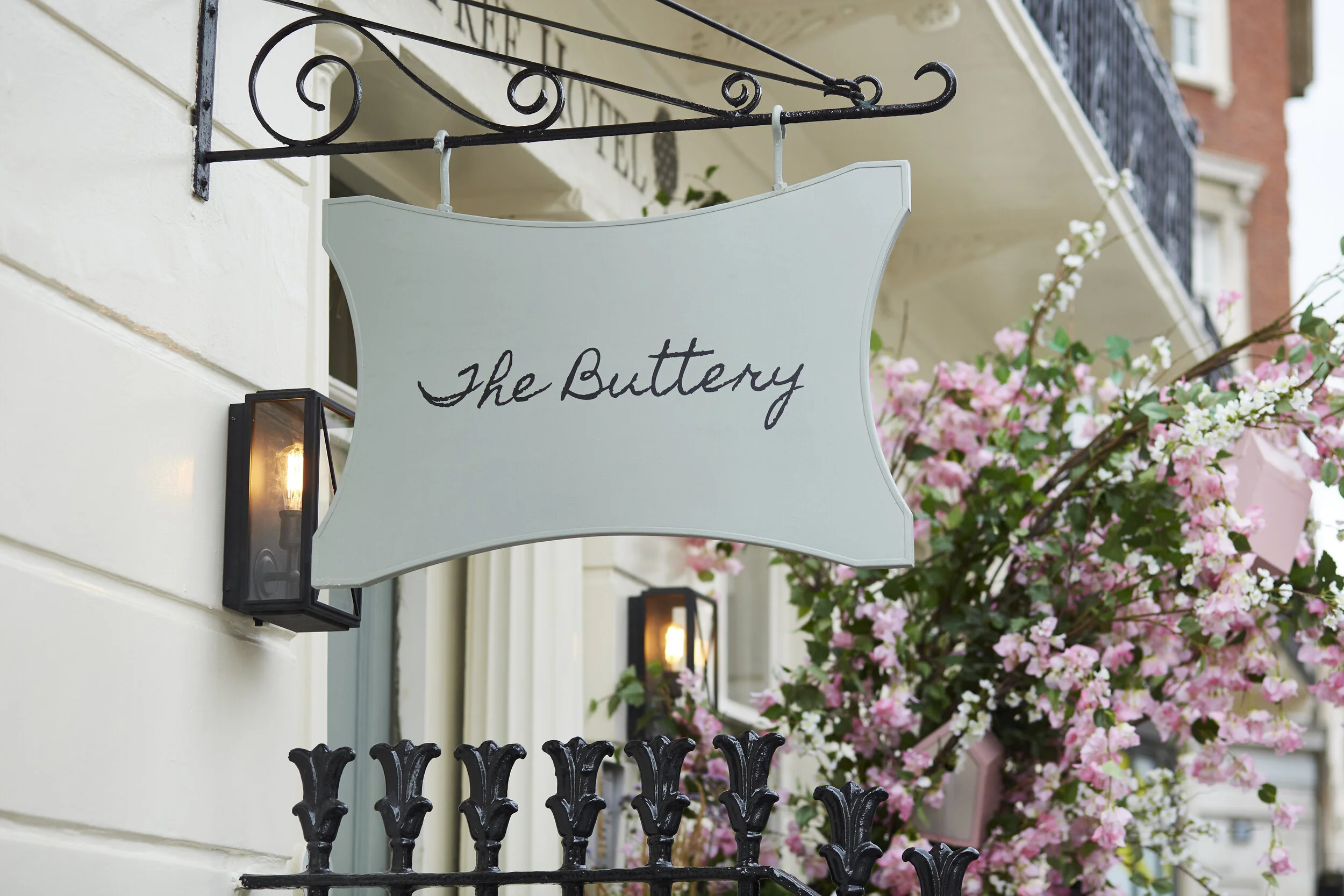 A sign reading 'The Buttery' hanging outside a building with orange flowers and black ornate fencing nearby.