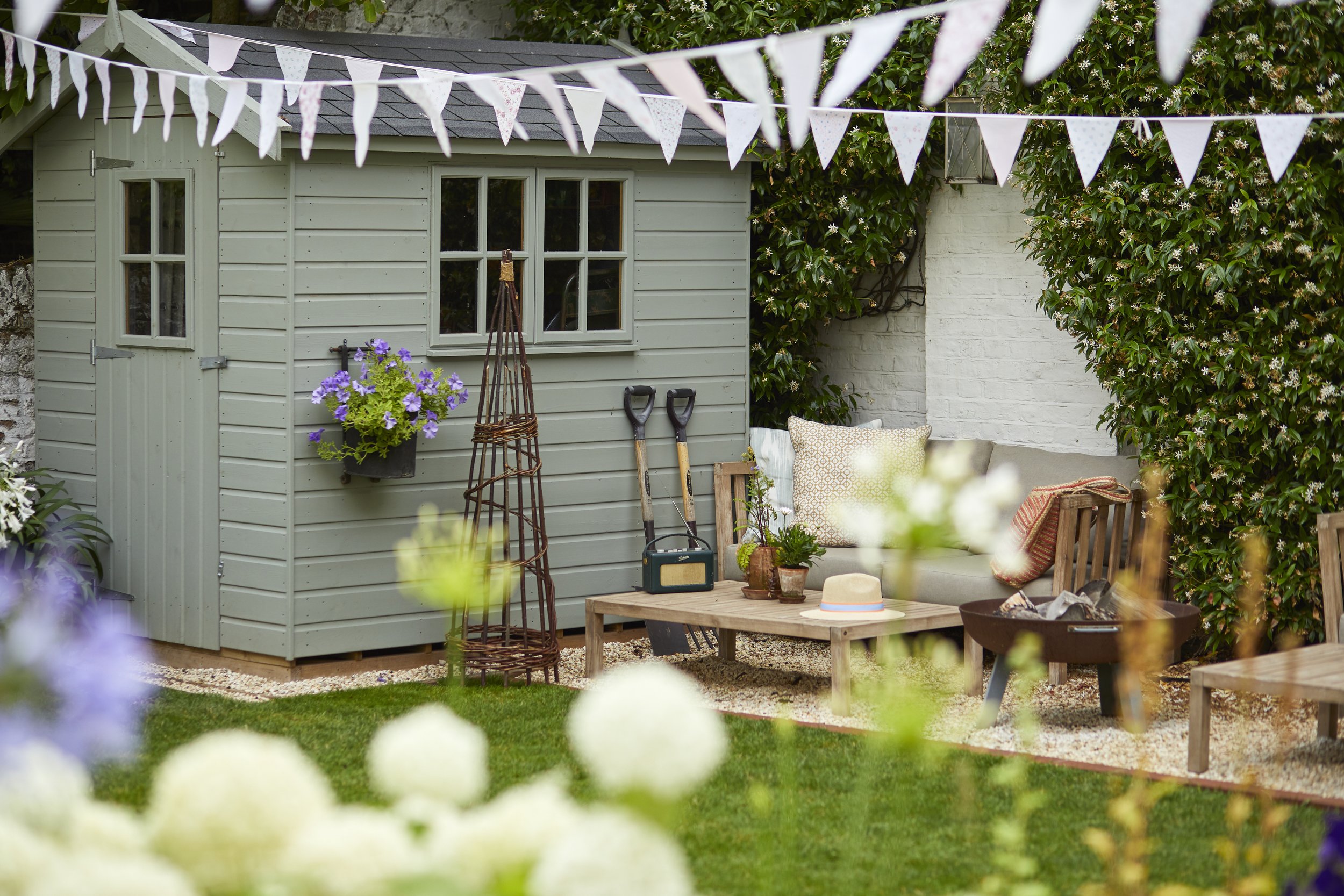 A backyard patio with a small green shed, a gray outdoor sofa with pillows, a wooden coffee table, potted plants, gardening tools, a fire pit, and bunting decorations hanging overhead. The area is surrounded by green bushes and flowers.