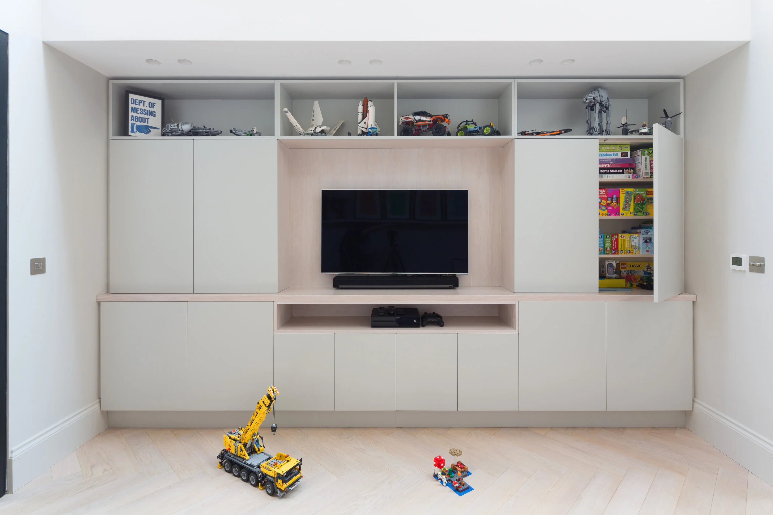 Modern living room with a built-in white entertainment center, a flat-screen TV, and shelves with toys including Lego vehicles and a small Lego figure, some books, and a yellow Lego crane on the floor.