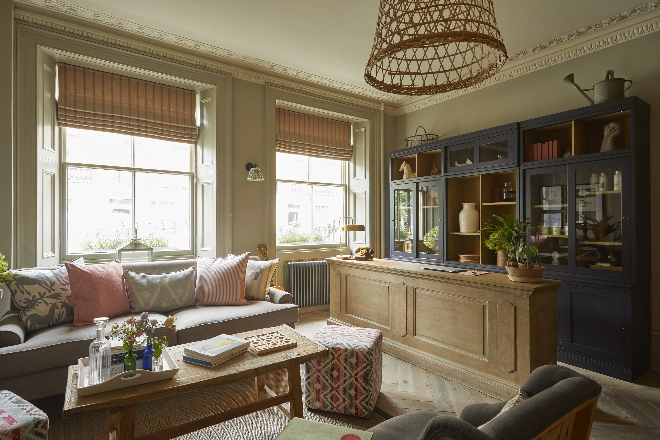 Cozy living room with large windows, beige walls, and decorative crown molding. A gray sofa with pink and patterned pillows, a wooden coffee table with glass bottles and books, a patterned ottoman, and a dark wood cabinet with glass doors displaying 