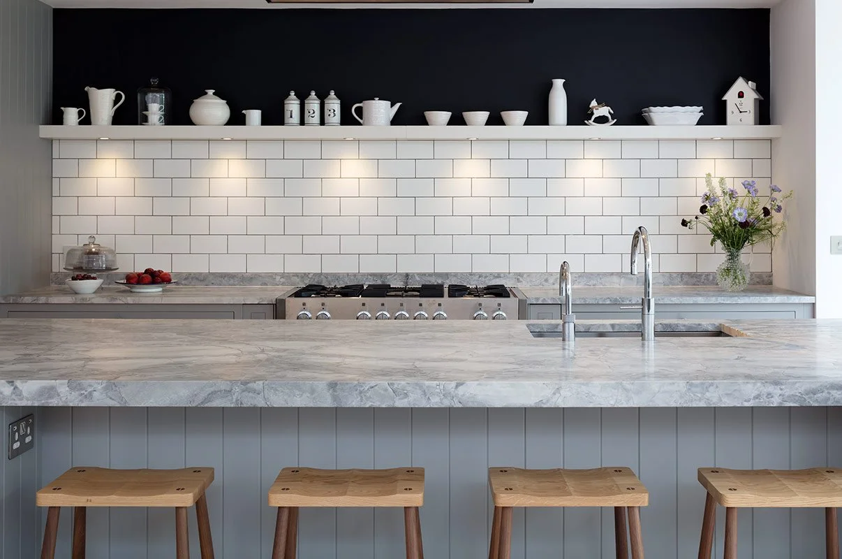 Modern kitchen with marble countertop island, white subway tile backsplash, open shelf with white dishware, stainless steel stove, and bar stools.