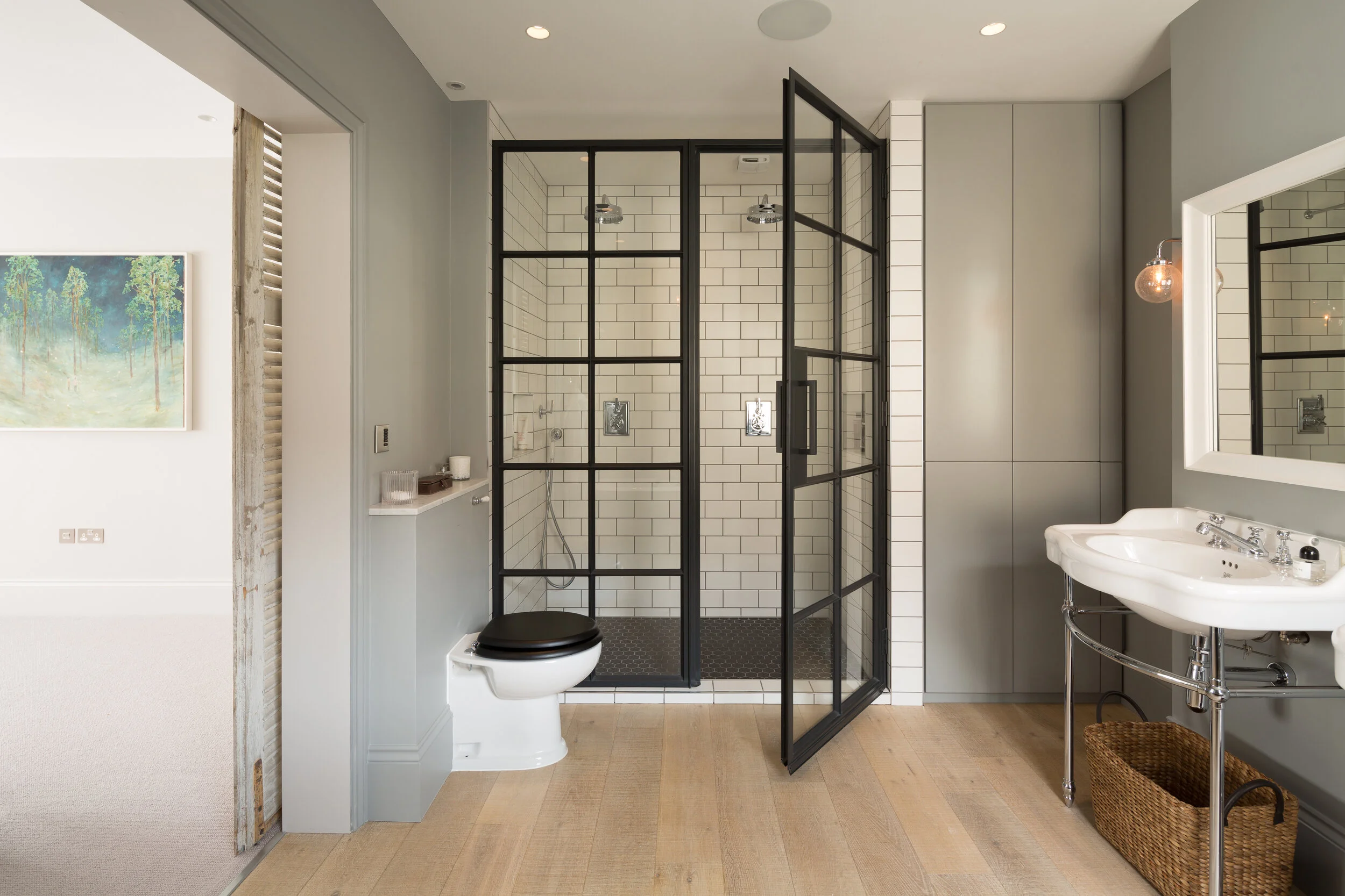 Modern bathroom with a glass-enclosed shower, toilet with black seat, white sink with chrome fixtures, large mirror with light, and a wicker basket on wooden flooring.