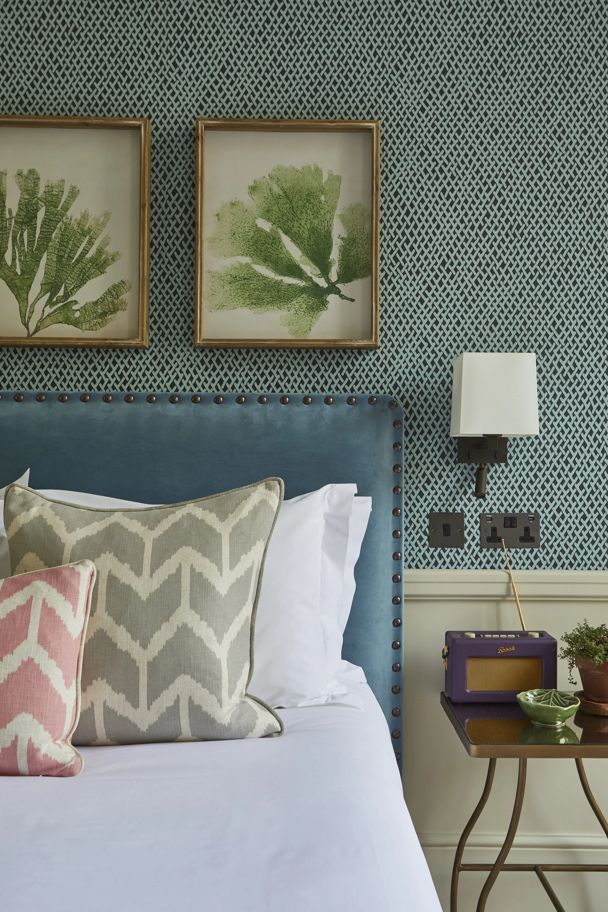 A bedroom with a blue upholstered headboard, decorative pillows in pink and beige with geometric patterns, two framed botanical prints on patterned wall, white wall sconce, purple radio on side table, and plants.