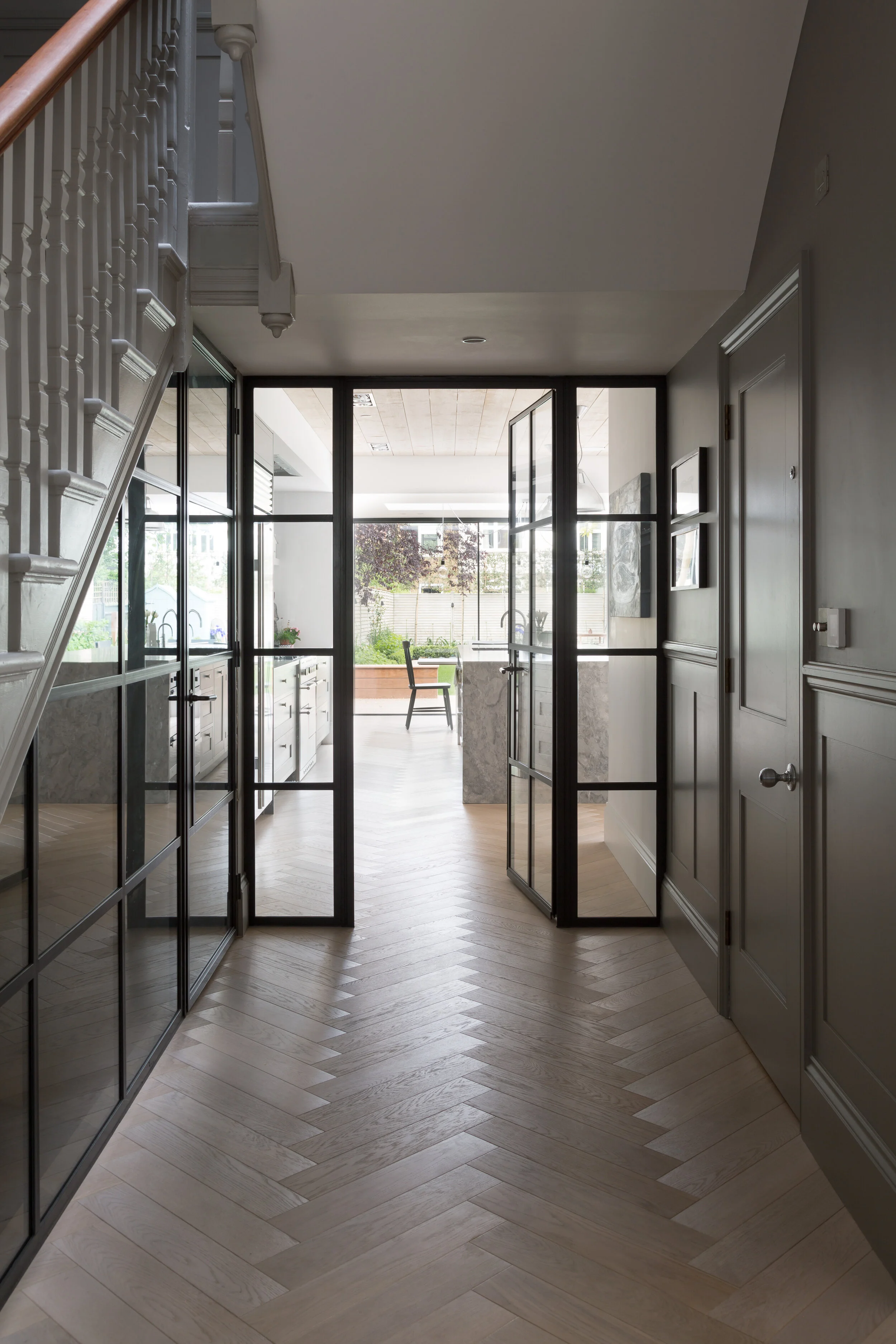 Modern kitchen area viewed through glass and metal door with light wood herringbone patterned floor.
