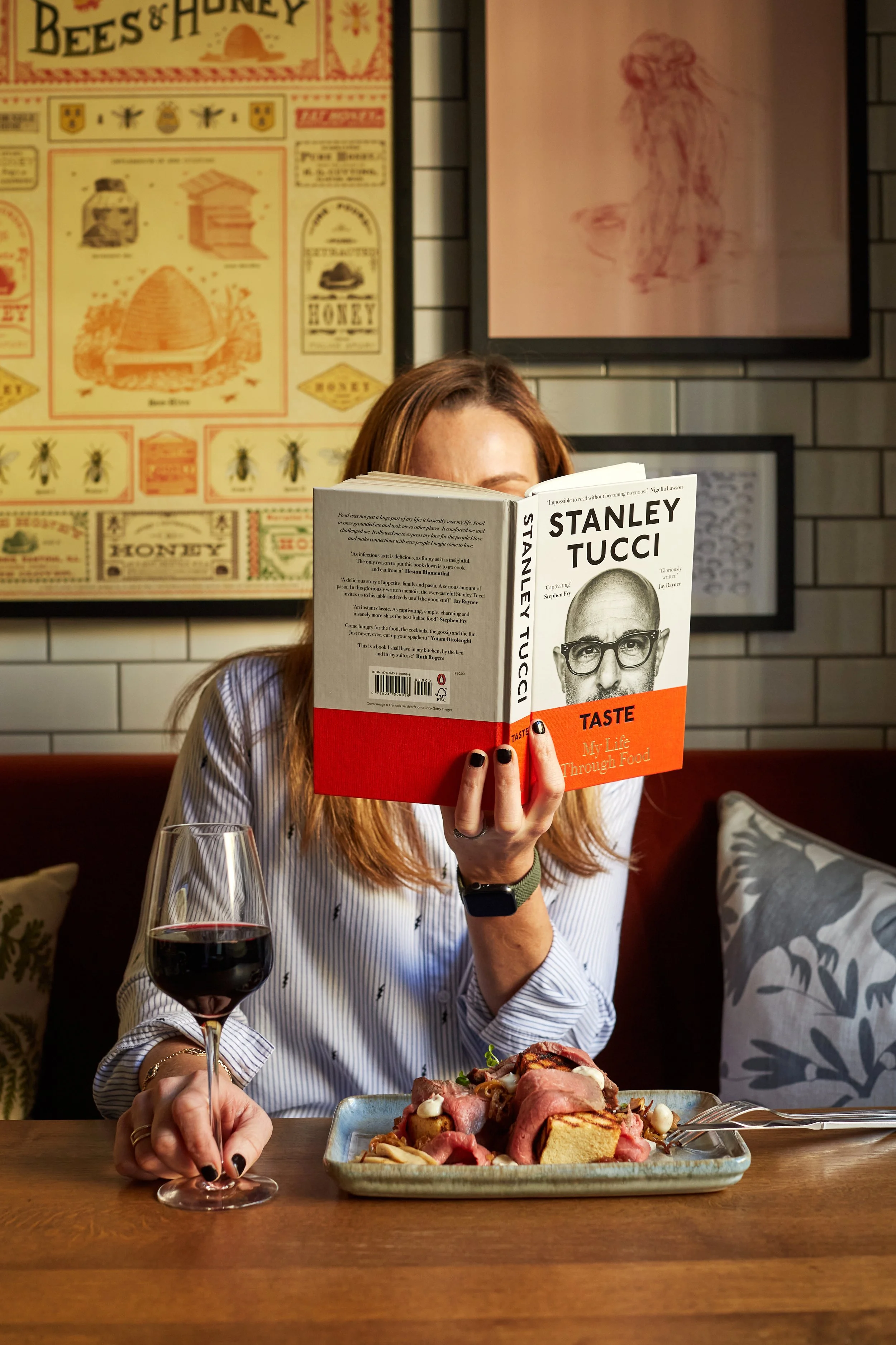 Woman with reddish hair reading a book titled 'Taste' by Stanley Tucci at a restaurant table with a glass of red wine and a plate of food, with posters and artwork on the wall behind her.