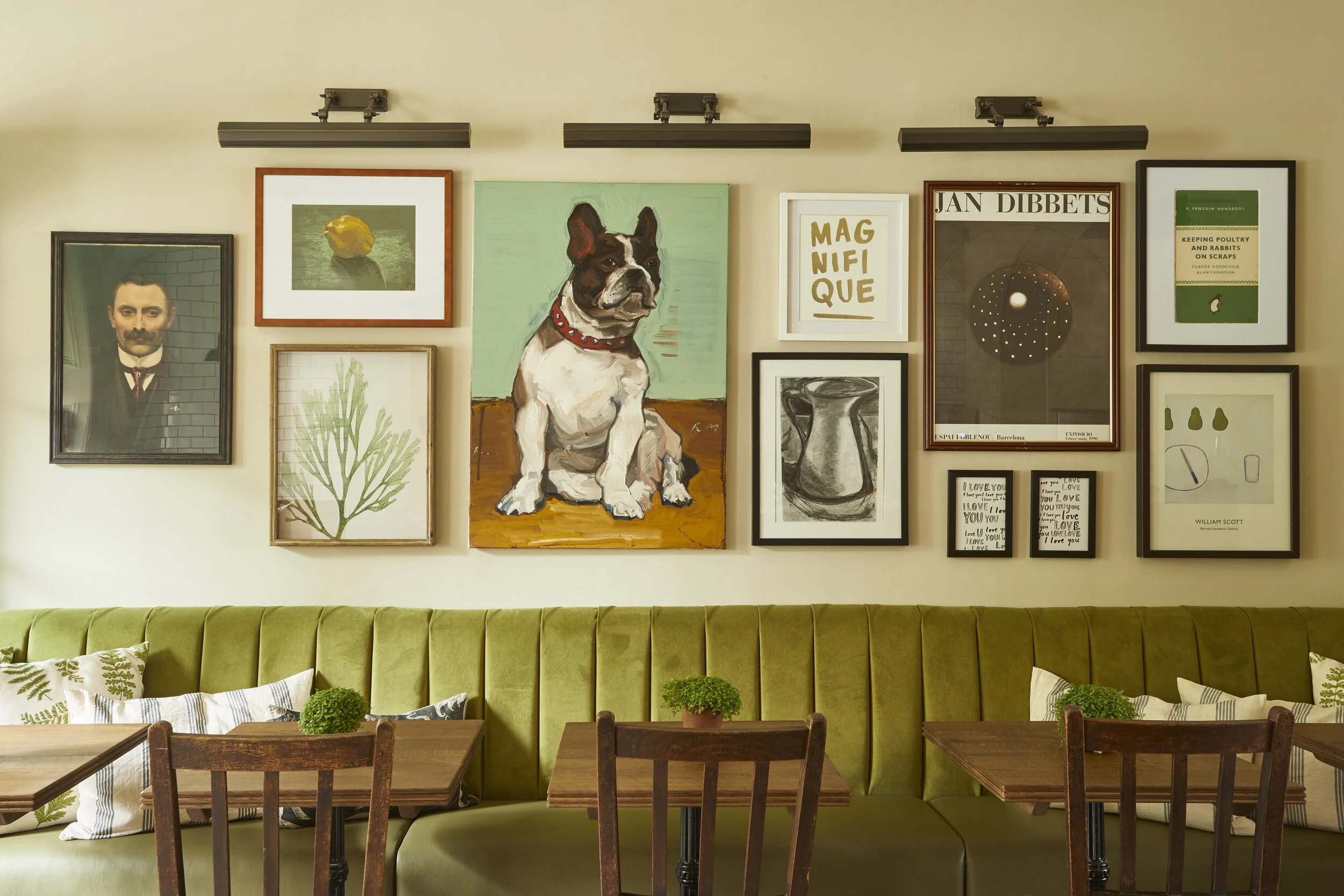 Wall decorated with various framed art pieces including a portrait of a man, a lemon, a dog painting, botanical print, book covers, and abstract images, with a green upholstered bench and wooden chairs and tables in the foreground.