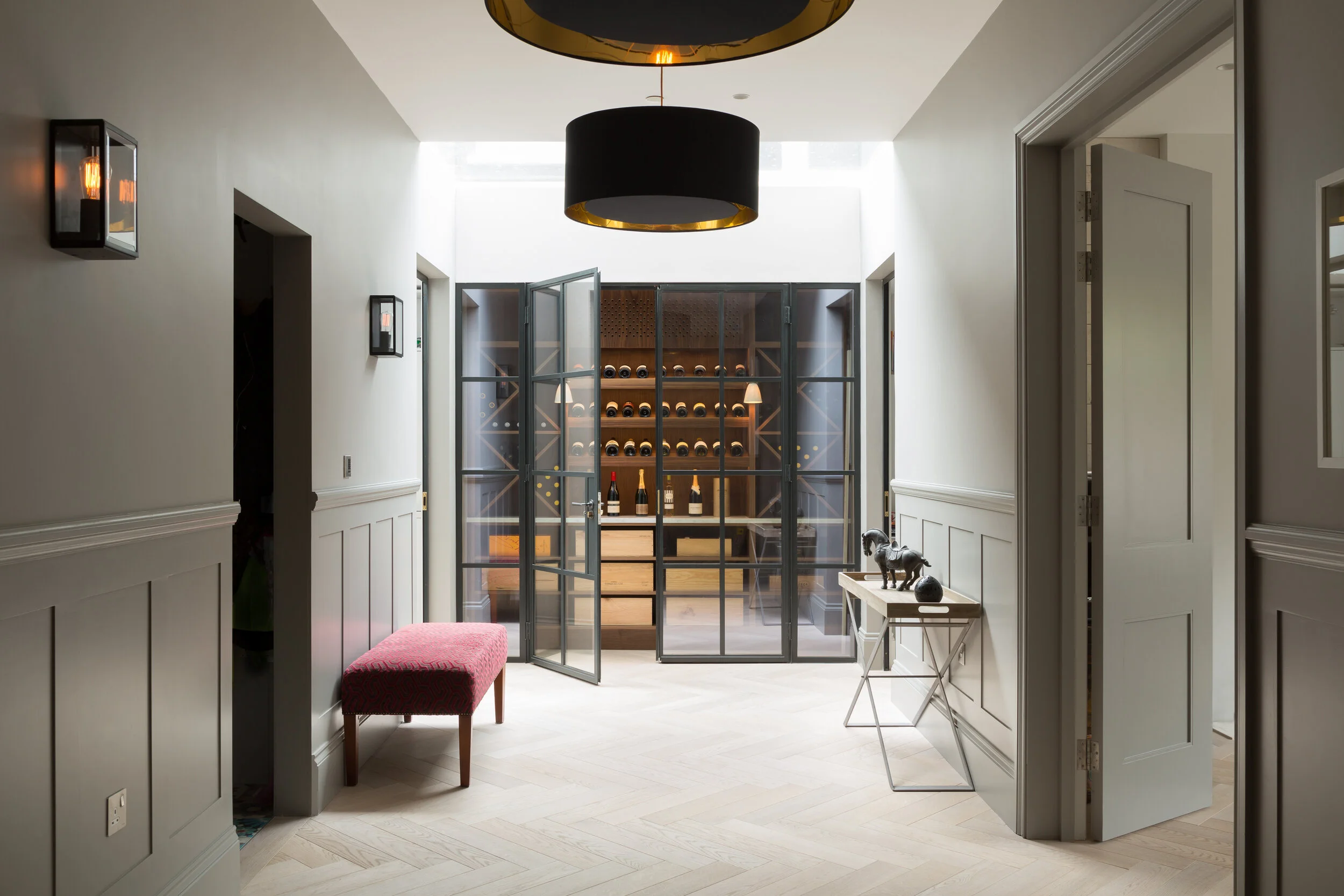 A stylish interior hallway with a glass door leading to a wine cellar, cream-colored walls with wainscoting, black pendant lights, a pink upholstered bench, and a side table with decorative objects.