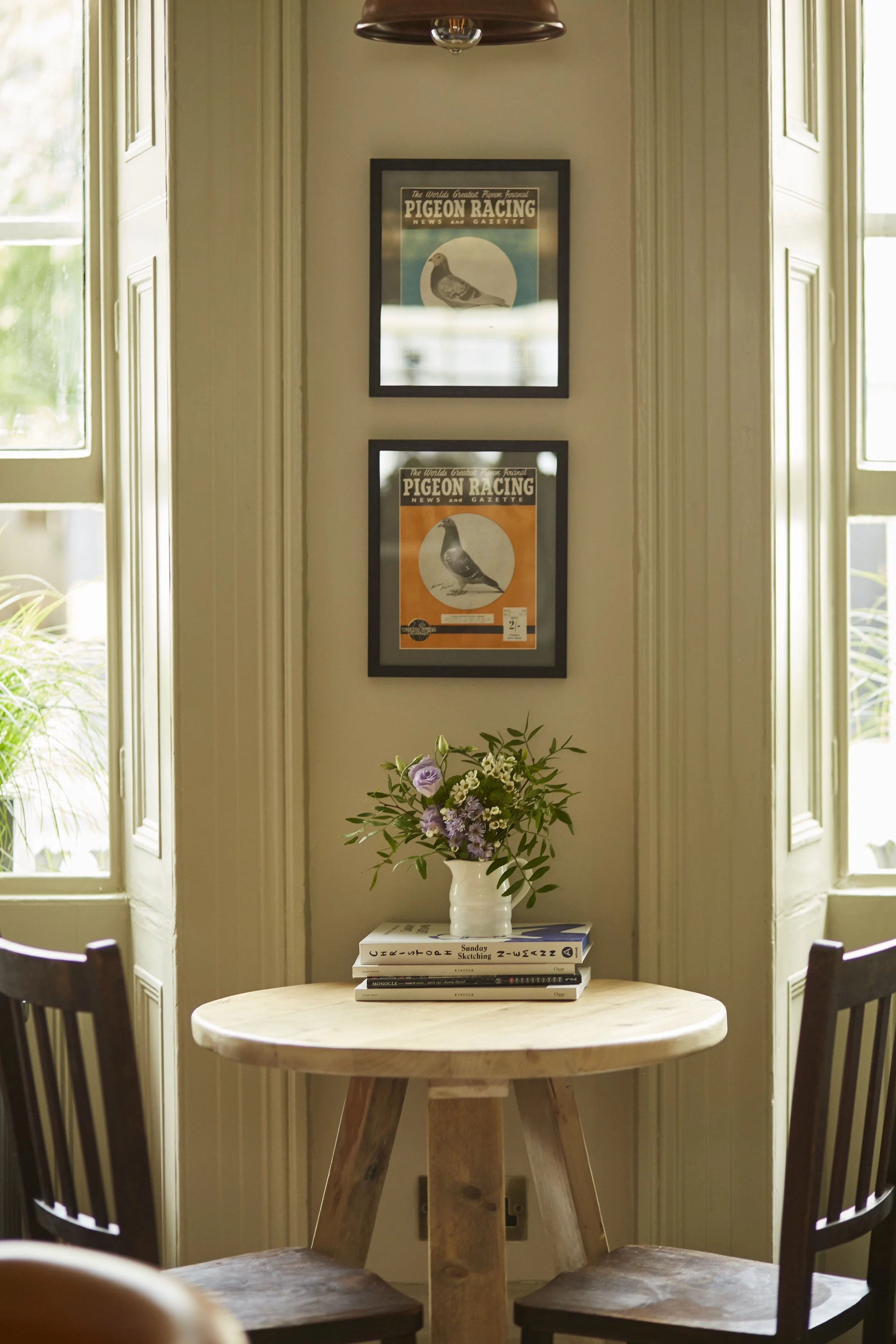 A cozy corner with a small round wooden table decorated with a white vase holding a bouquet of purple and green flowers, and four stacked books. Two framed pigeon racing magazines hang above the table, flanked by two large windows with white trim, al