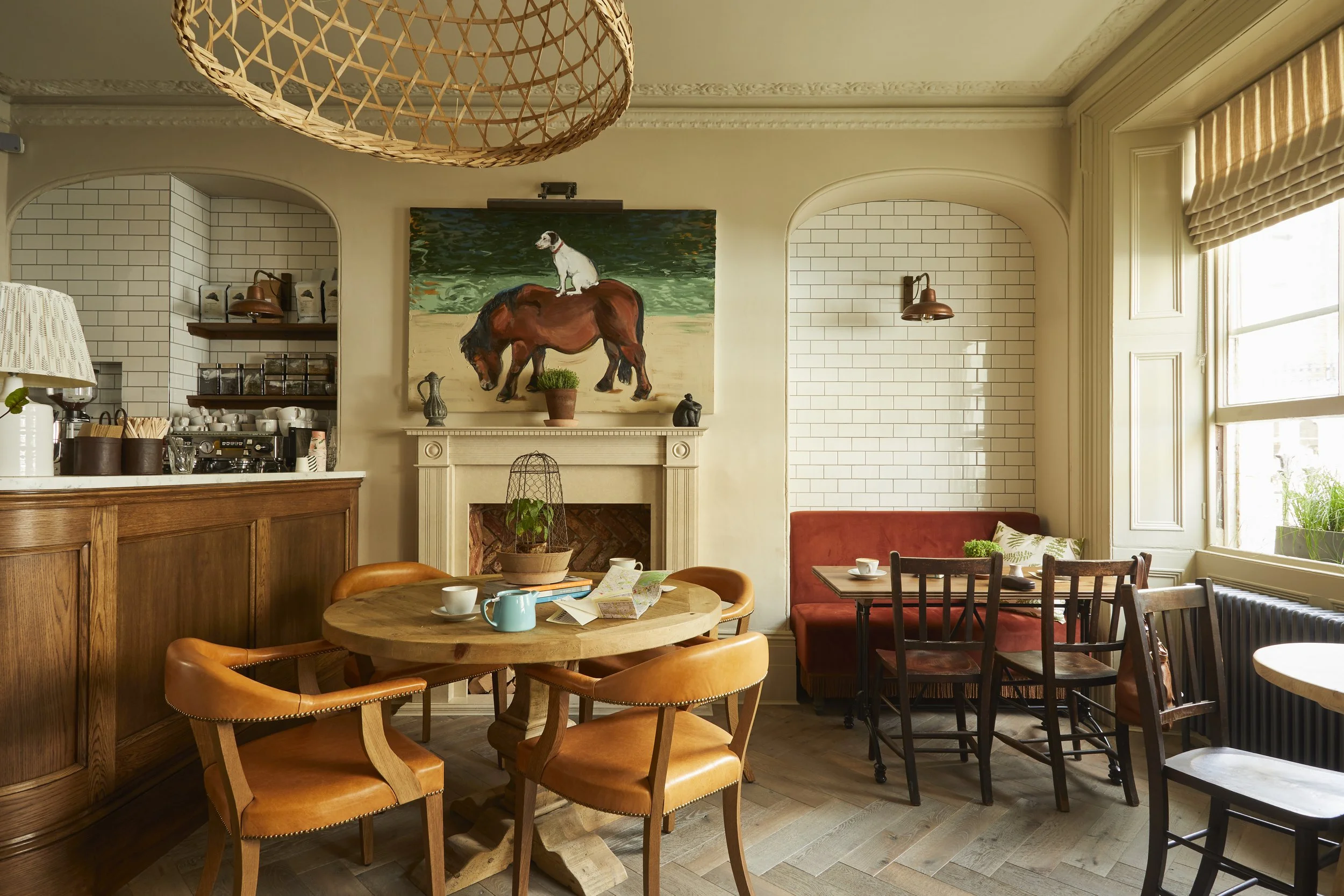 Interior of a cozy cafe with wooden tables and chairs, white brick walls, a fireplace with a painting of a dog on a horse above it, and large windows letting in natural light.