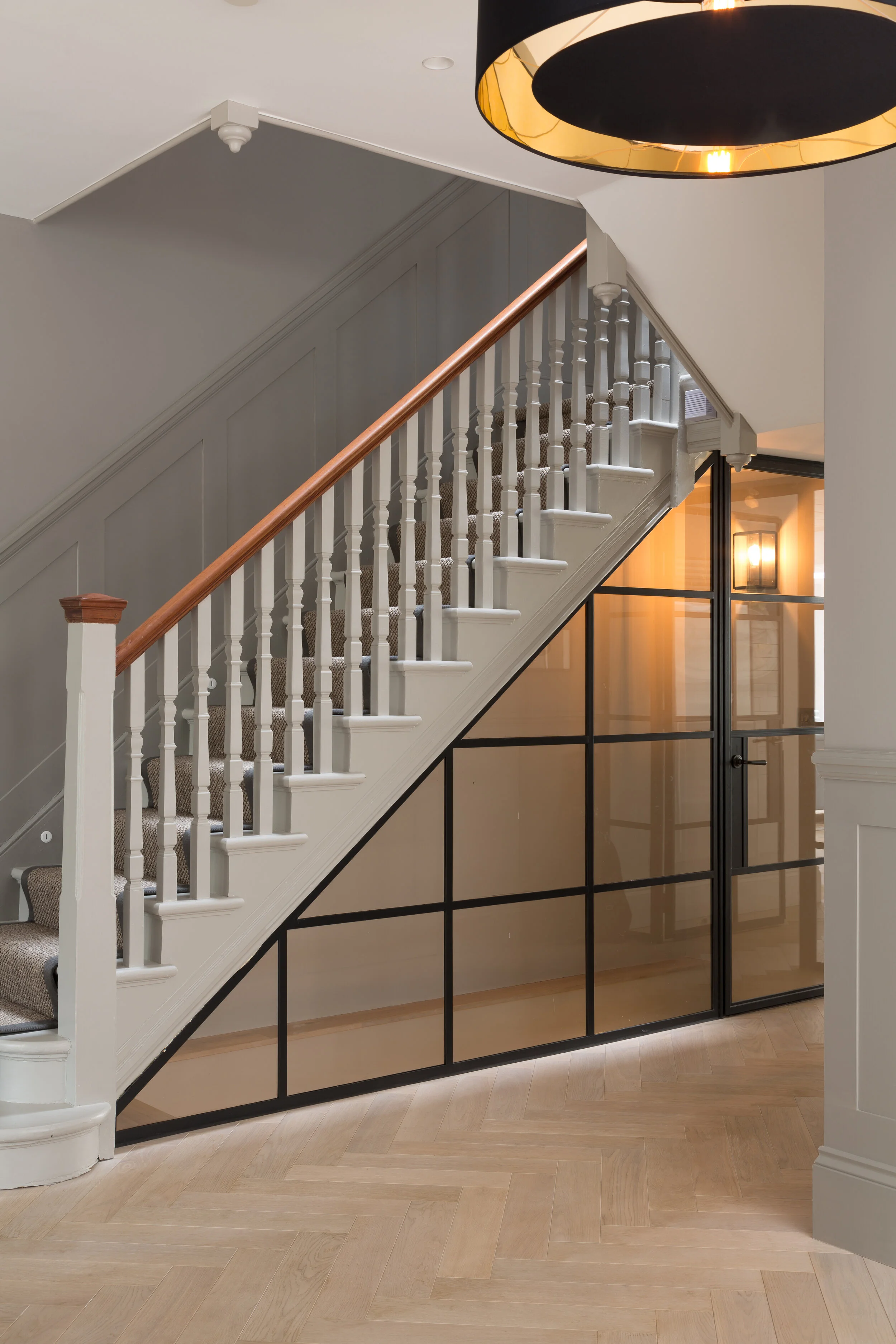Interior view of a staircase with white railing and brown handrail, black-framed glass partition underneath, wood herringbone floor, and modern ceiling and wall lighting.
