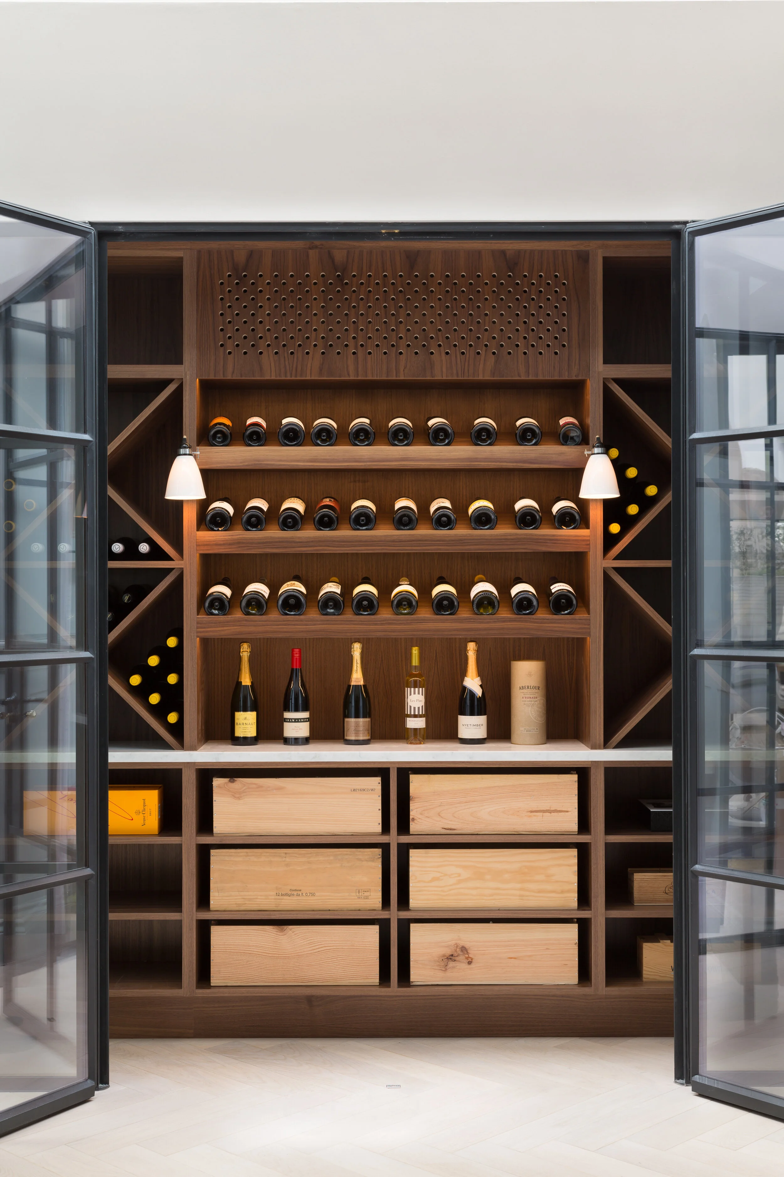 A wine cellar with wooden shelves holding multiple bottles of wine, bottles stored horizontally on the top three shelves and vertically in side compartments, with several bottles of champagne and wine on a middle shelf. There are also wooden boxes an
