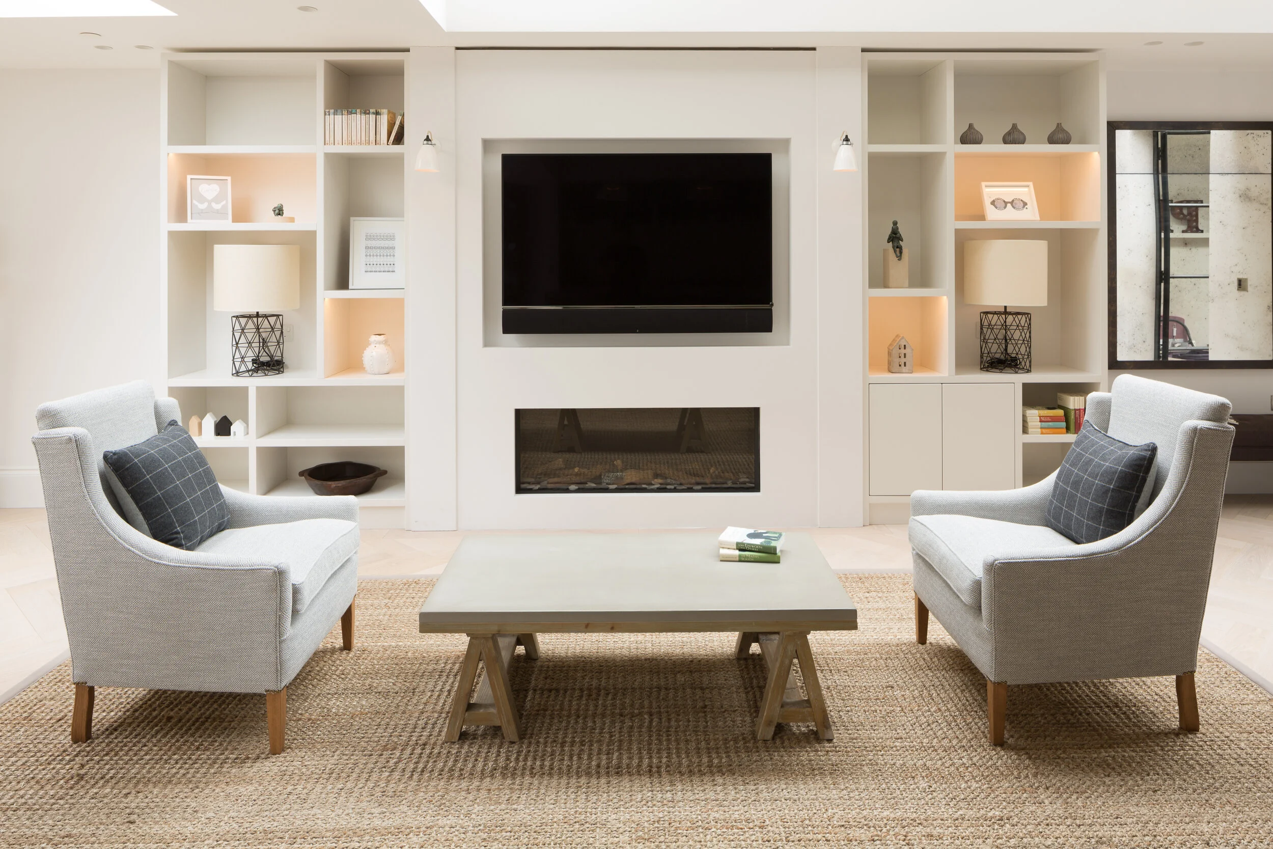 Living room with two light gray armchairs, a rectangular coffee table, white built-in shelves, a wall-mounted flat-screen TV, a modern fireplace, and decorative items on the shelves.