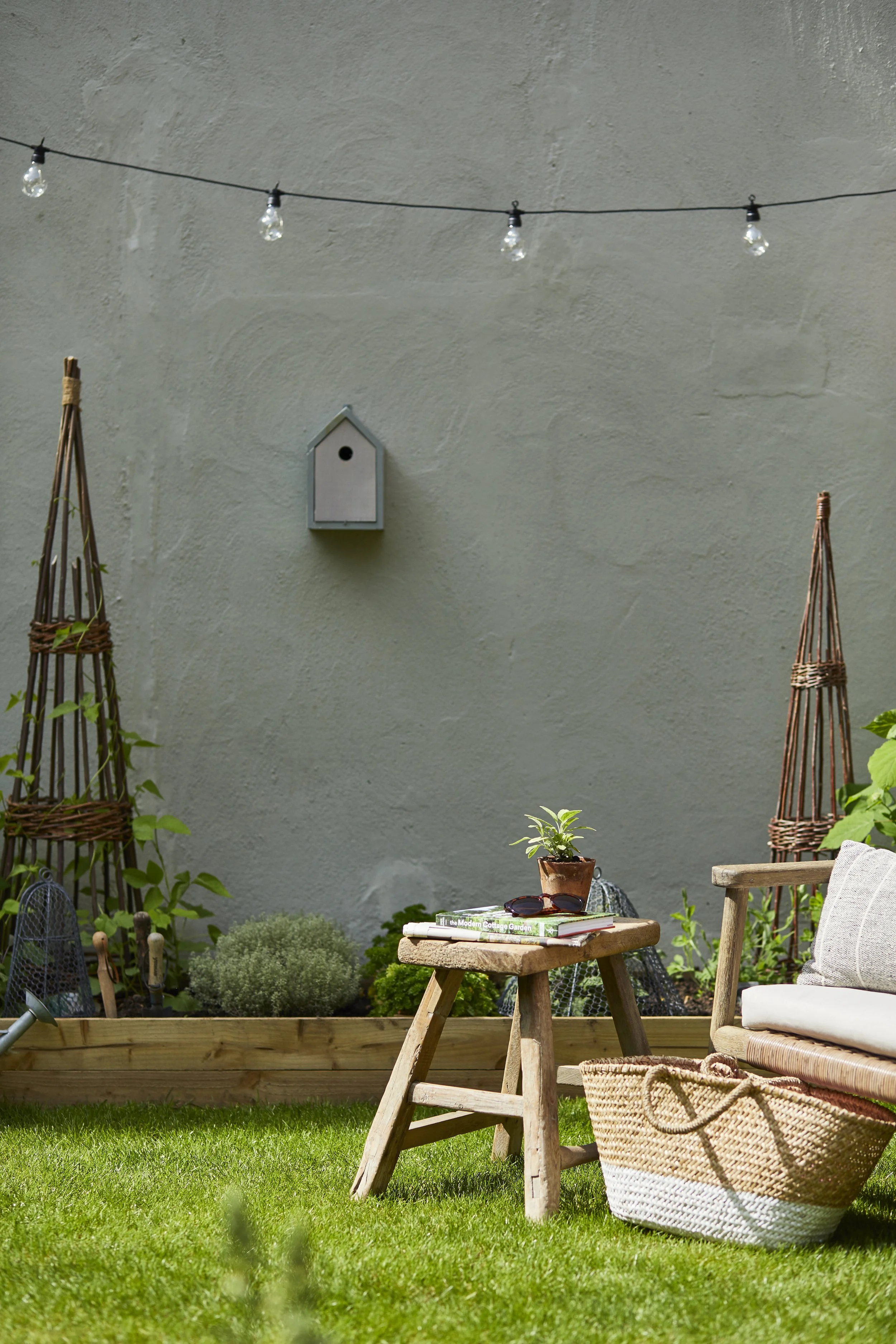 A cozy outdoor garden space with a gray wall, string lights, a small birdhouse, wooden trellises, plants, a wooden table with potted plant and sunglasses, and a woven basket next to a bench with cushion.