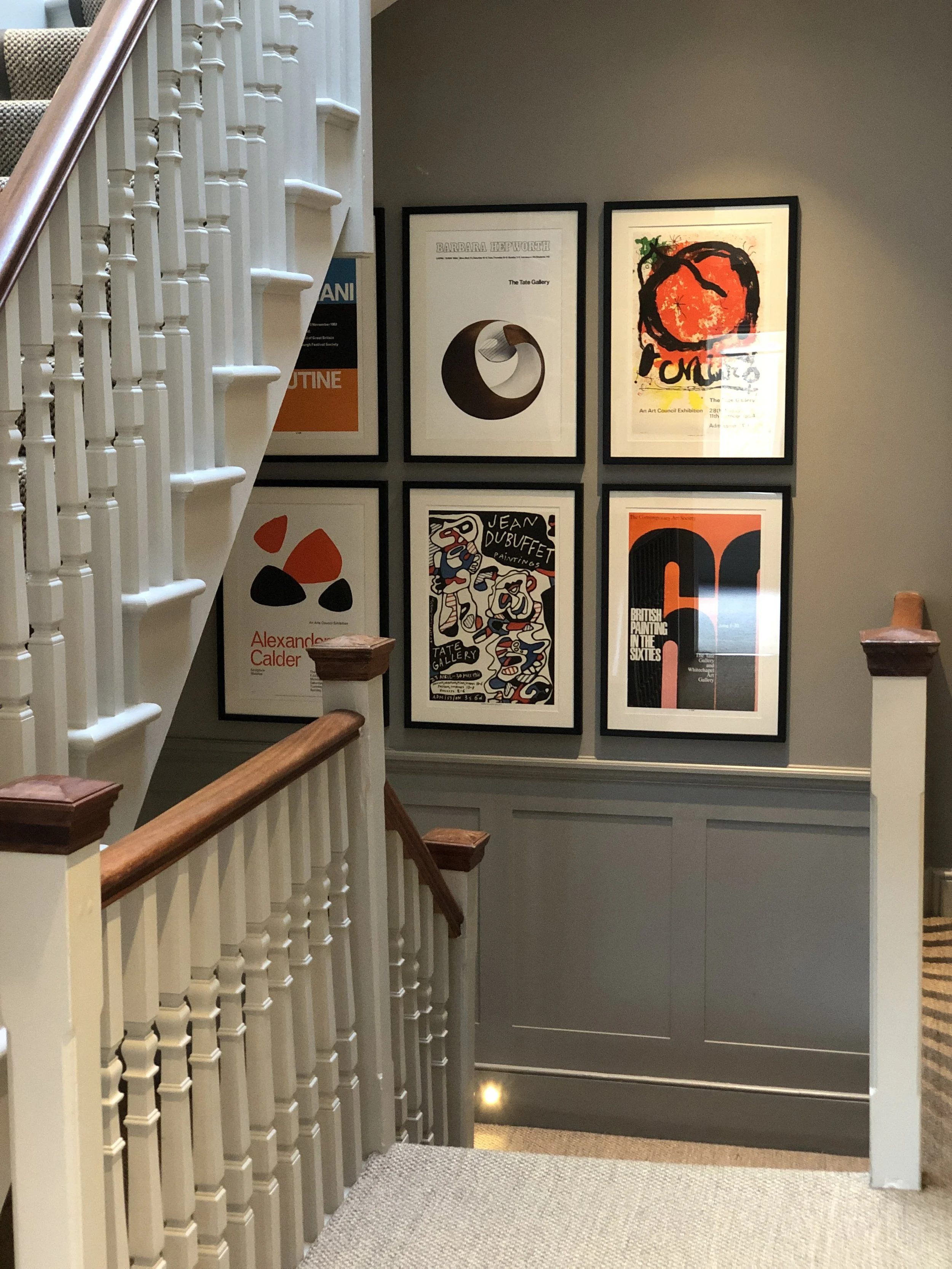 Gallery staircase landing with framed art posters on a gray wall, featuring abstract and modern artwork, with white painted balustrades and wooden handrails.