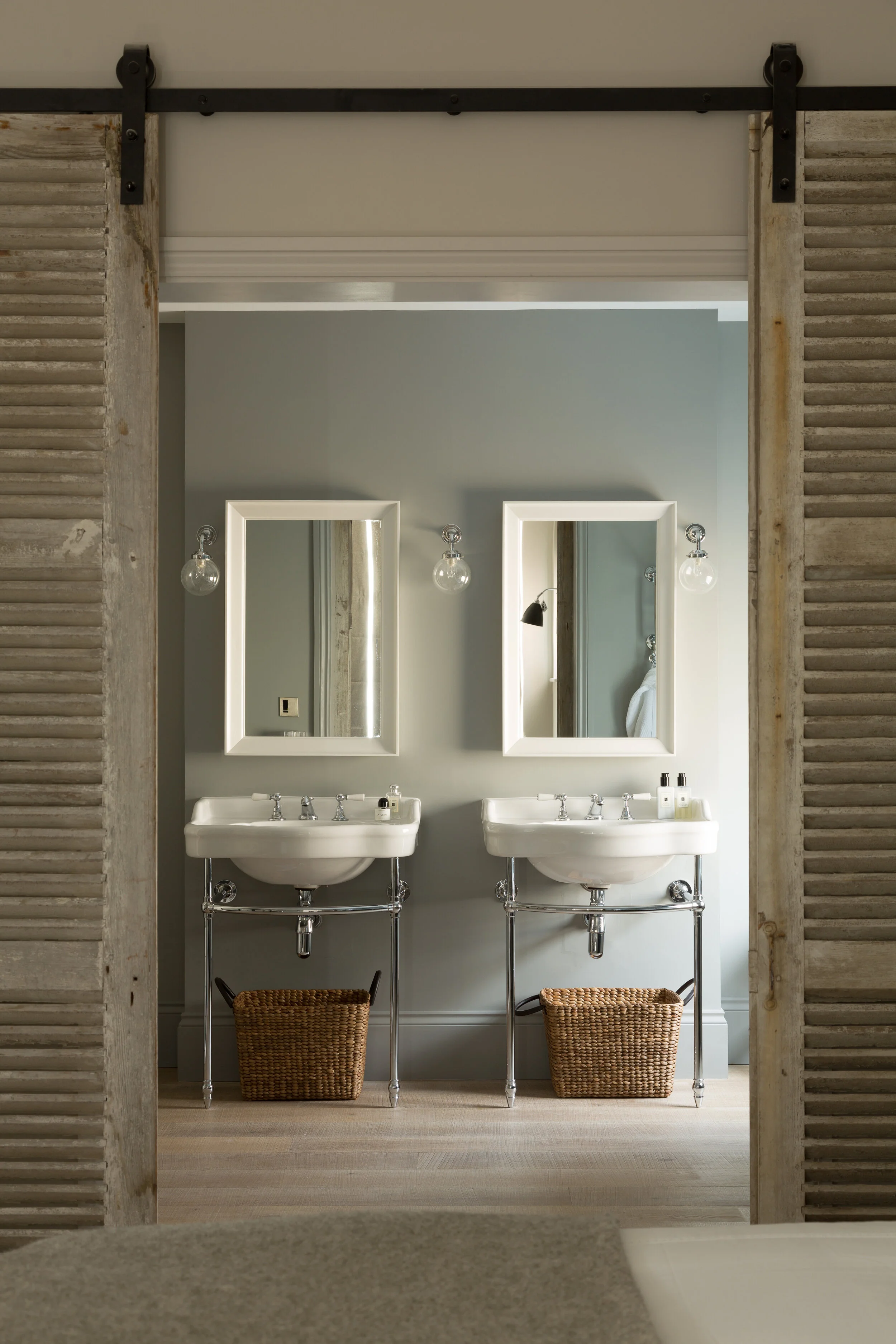 An interior view of a bathroom with two white sinks, each with a mirror above, two light bulbs on either side, and baskets underneath, seen through rustic wooden sliding barn doors.