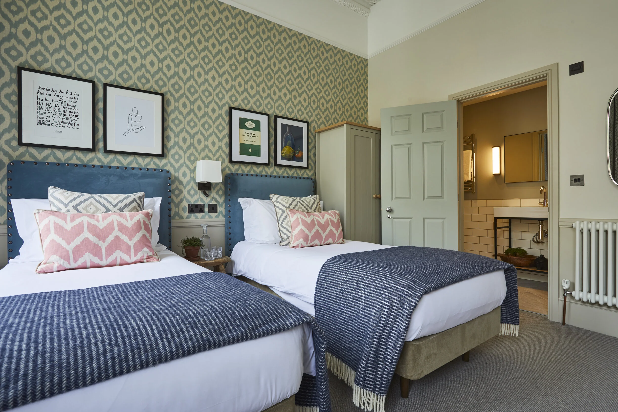 A hotel room with two beds, blue upholstered headboards, patterned and plain pillows, bedside lamp, framed artwork above the beds, and an open door leading to a bathroom with a sink and mirror.