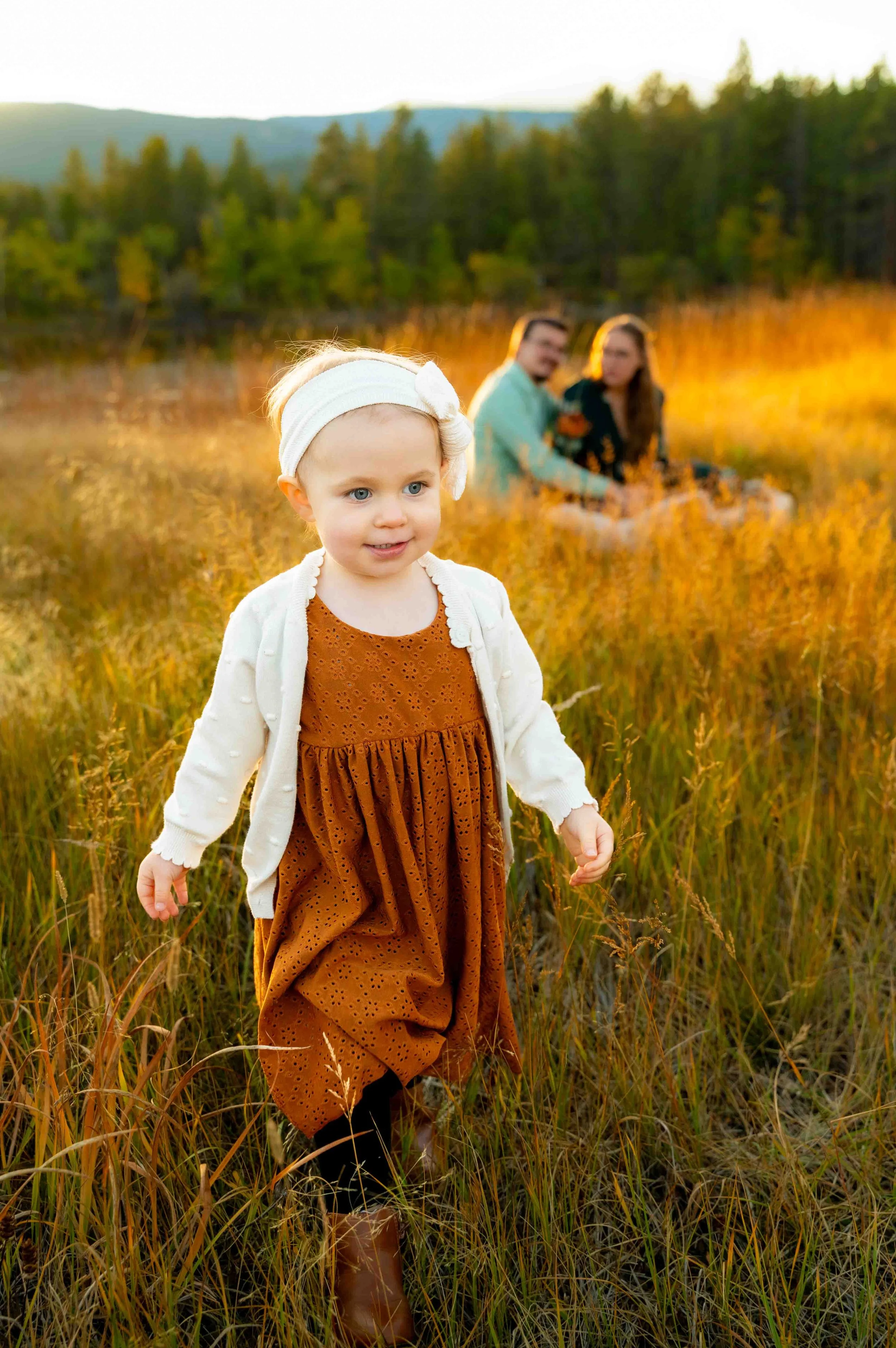 Parents sit in the grass while their daughter explores the meadow during their family photoshoot in the Rocky Mountains