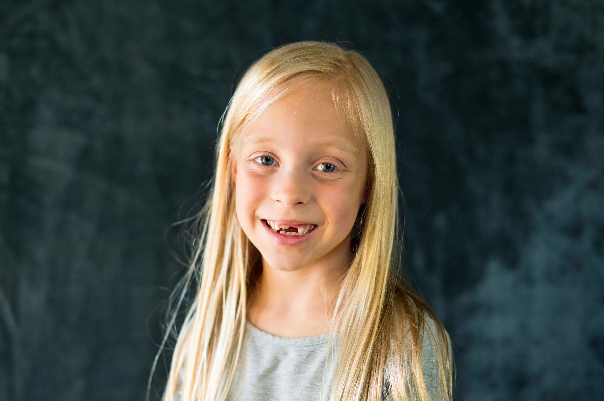 Girl with blonde hair smiles at the camera with two missing front teeth during her session with the Longmont school photographer, Sara Herkes