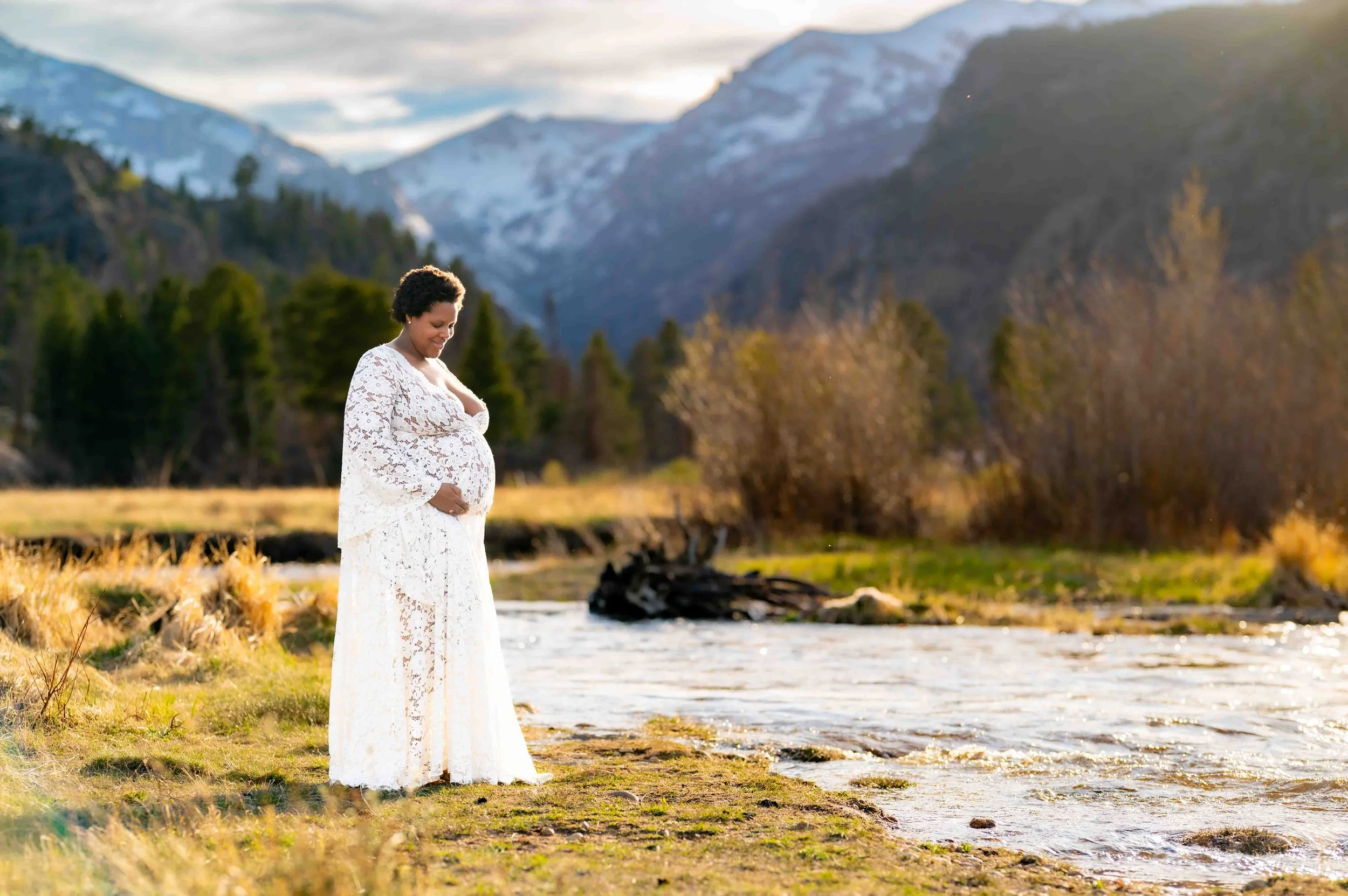 Pregnant woman stands looking at her belly near a stream with towering mountains behind during her Rocky Mountain National Park Maternity Session