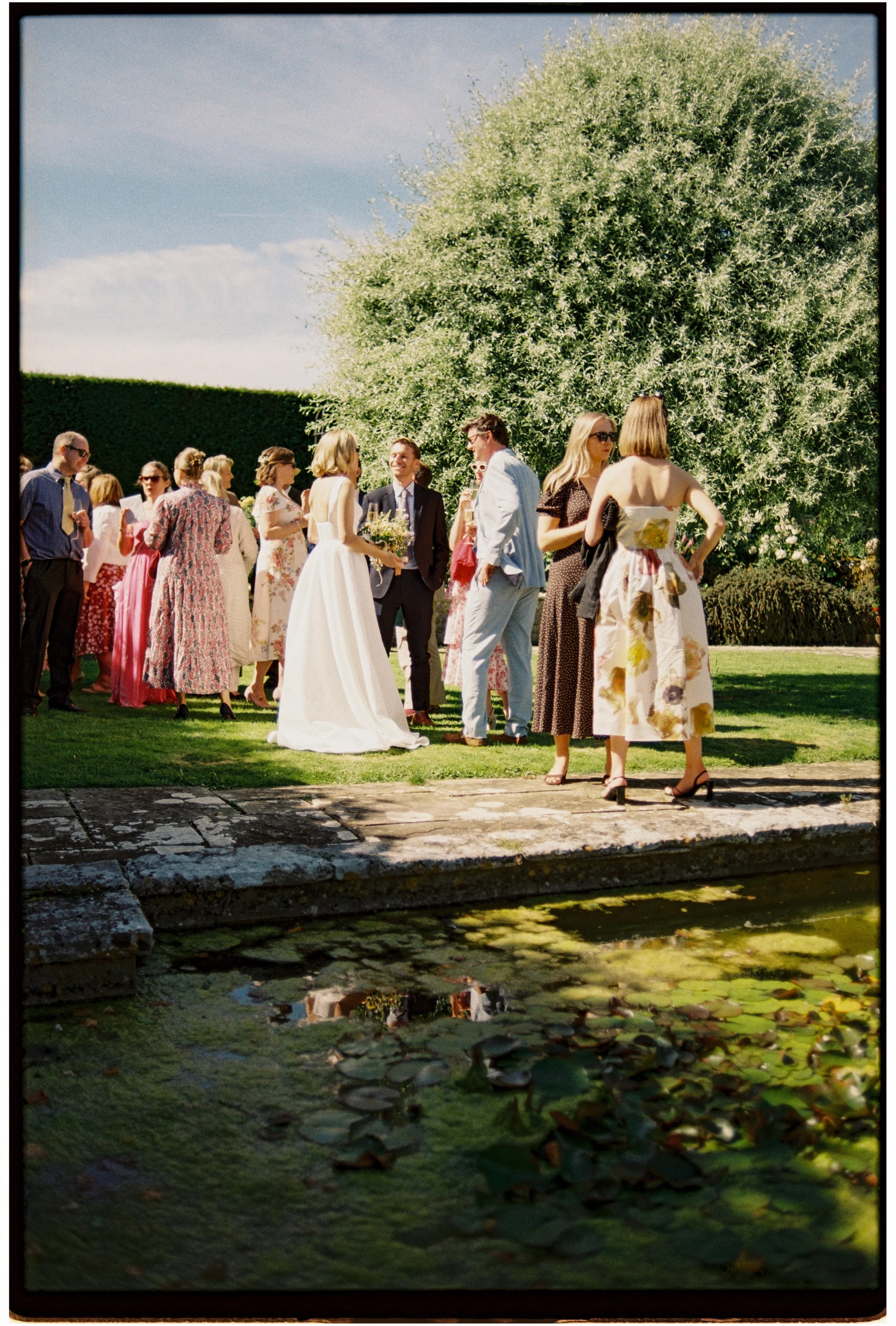A group of people gathered outdoors near a large tree on a sunny day, with some socializing and chatting, while a woman in a white dress holds a bouquet.