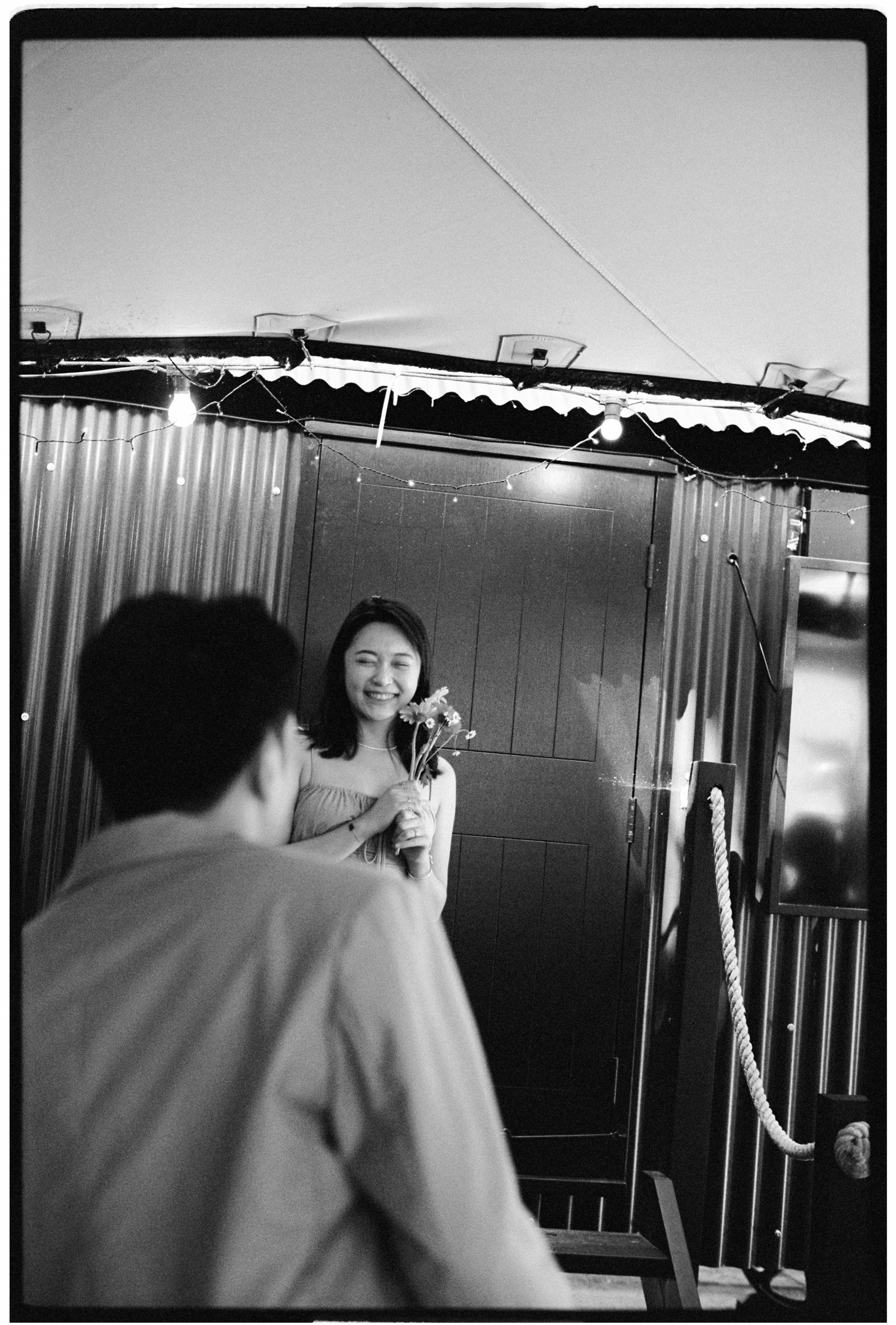 A woman smiling and holding flowers, with a man in the foreground, in a room with string lights and wooden paneling.