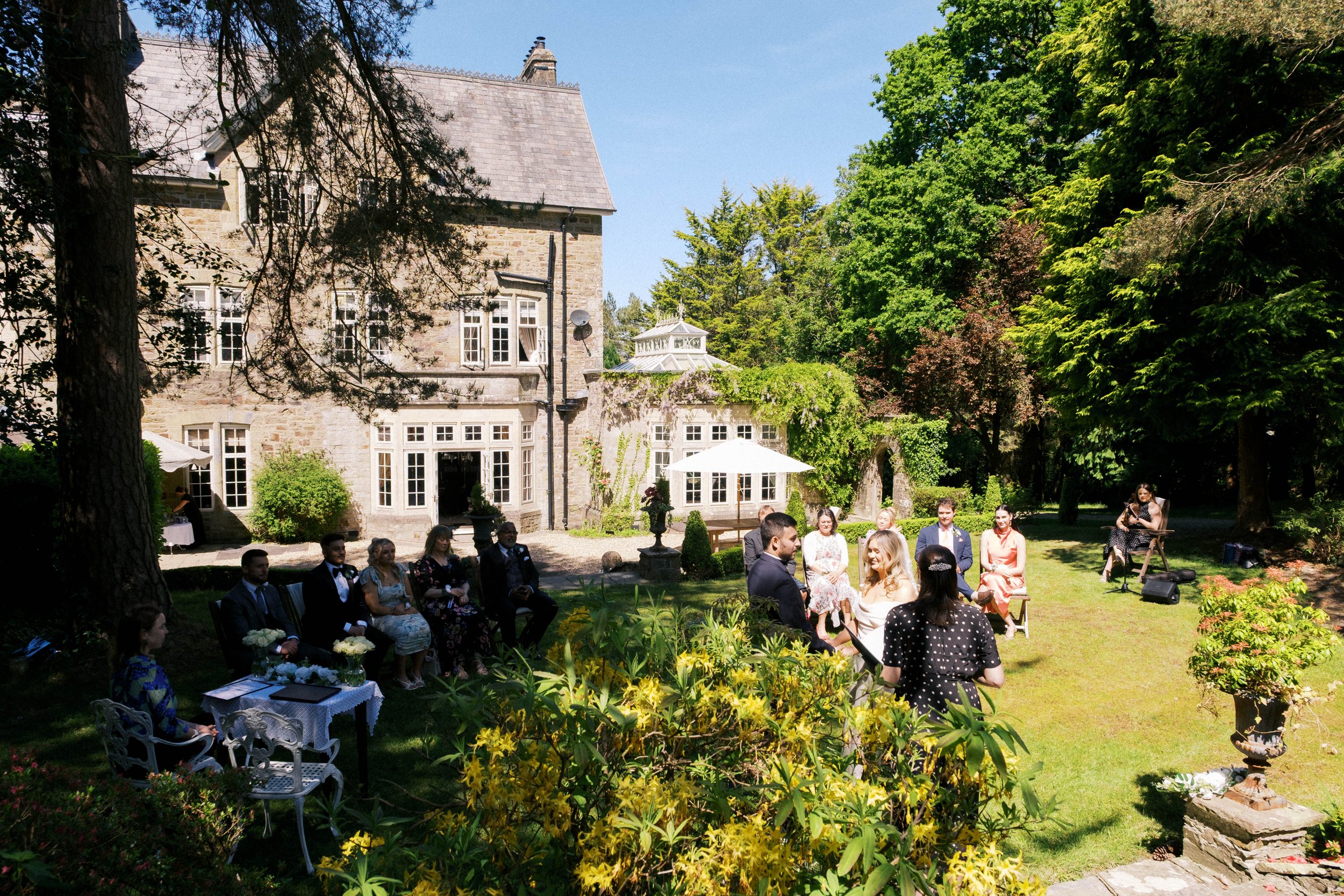 People celebrating at a wedding outdoors in a garden with a large stone house and lush trees in the background.