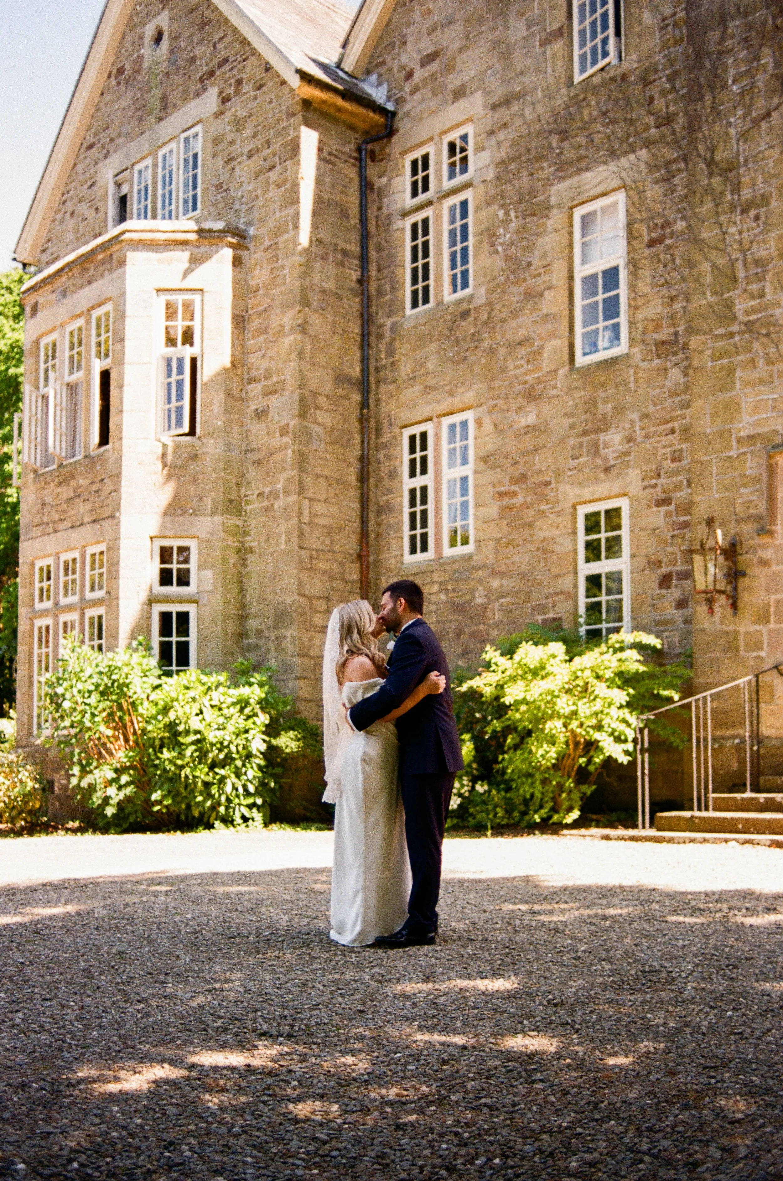 A bride and groom standing close together and kissing outdoors in front of a large stone building with multiple windows. The bride is wearing a white dress and veil, and the groom is in a black suit. There are green bushes and a staircase nearby, wit