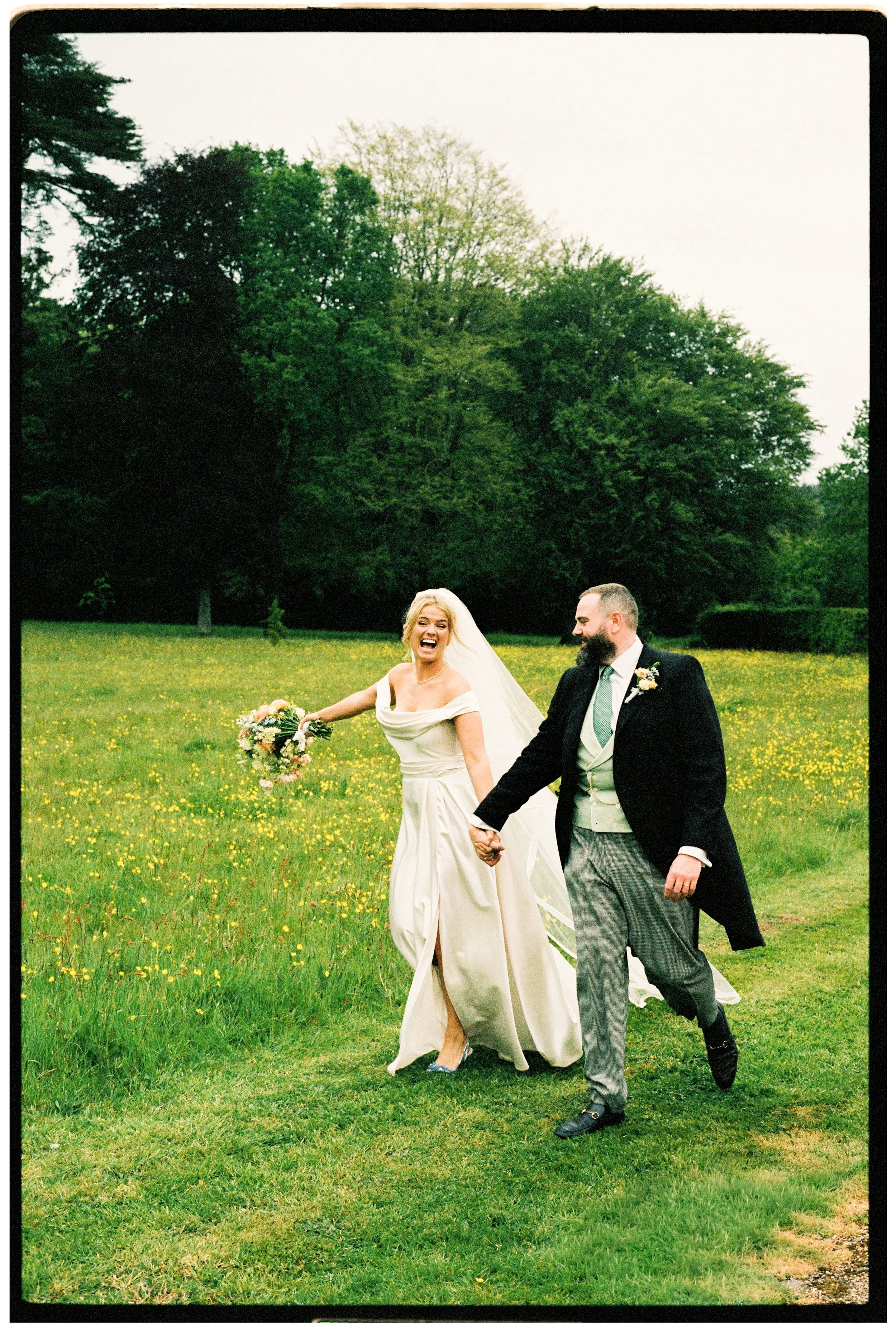 A bride and groom holding hands and smiling while walking through a green field with yellow flowers, trees in the background, and an overcast sky.