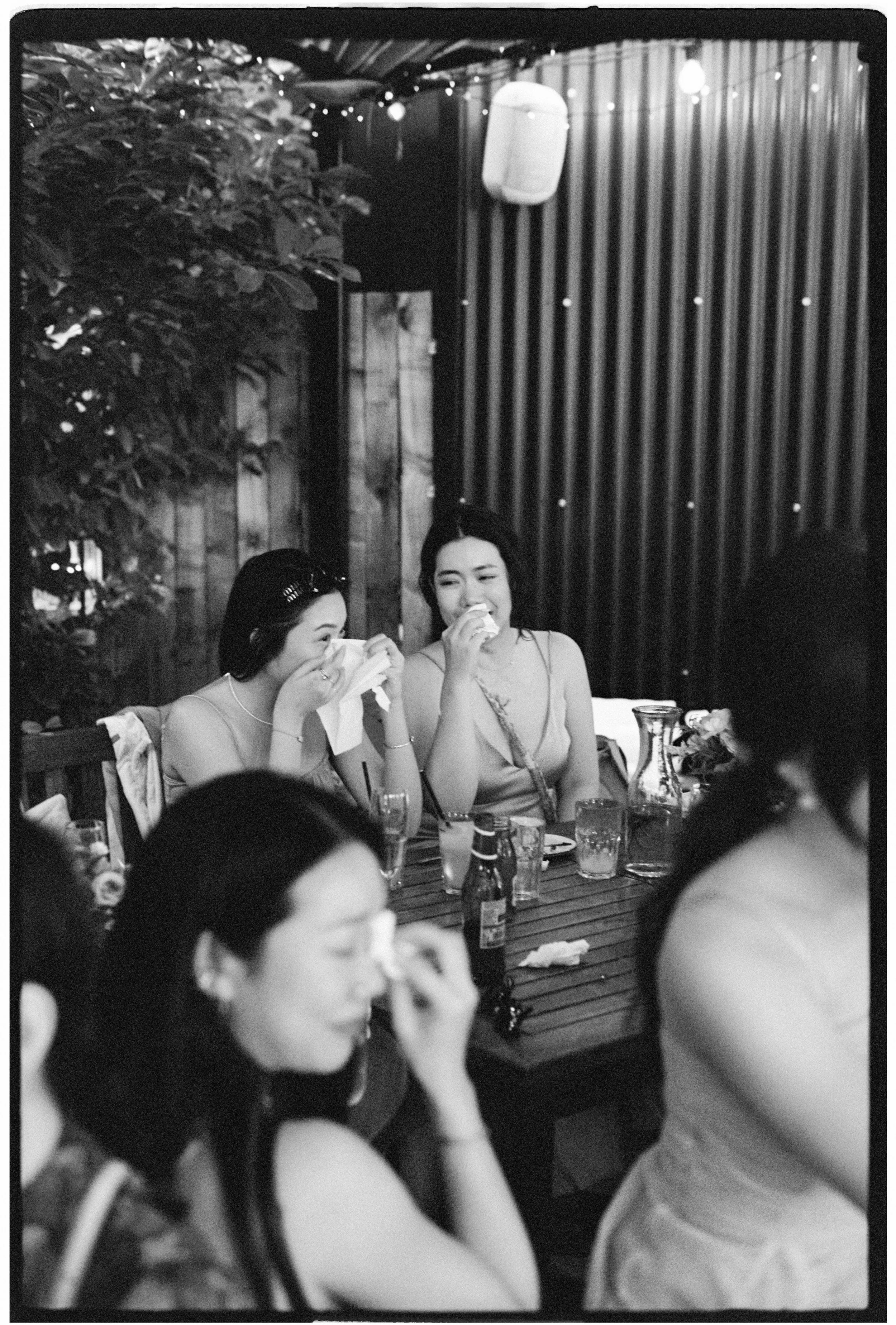 Black and white photo of women sitting at a table, some drinking, in a cozy, decorated room with string lights and a wooden wall.