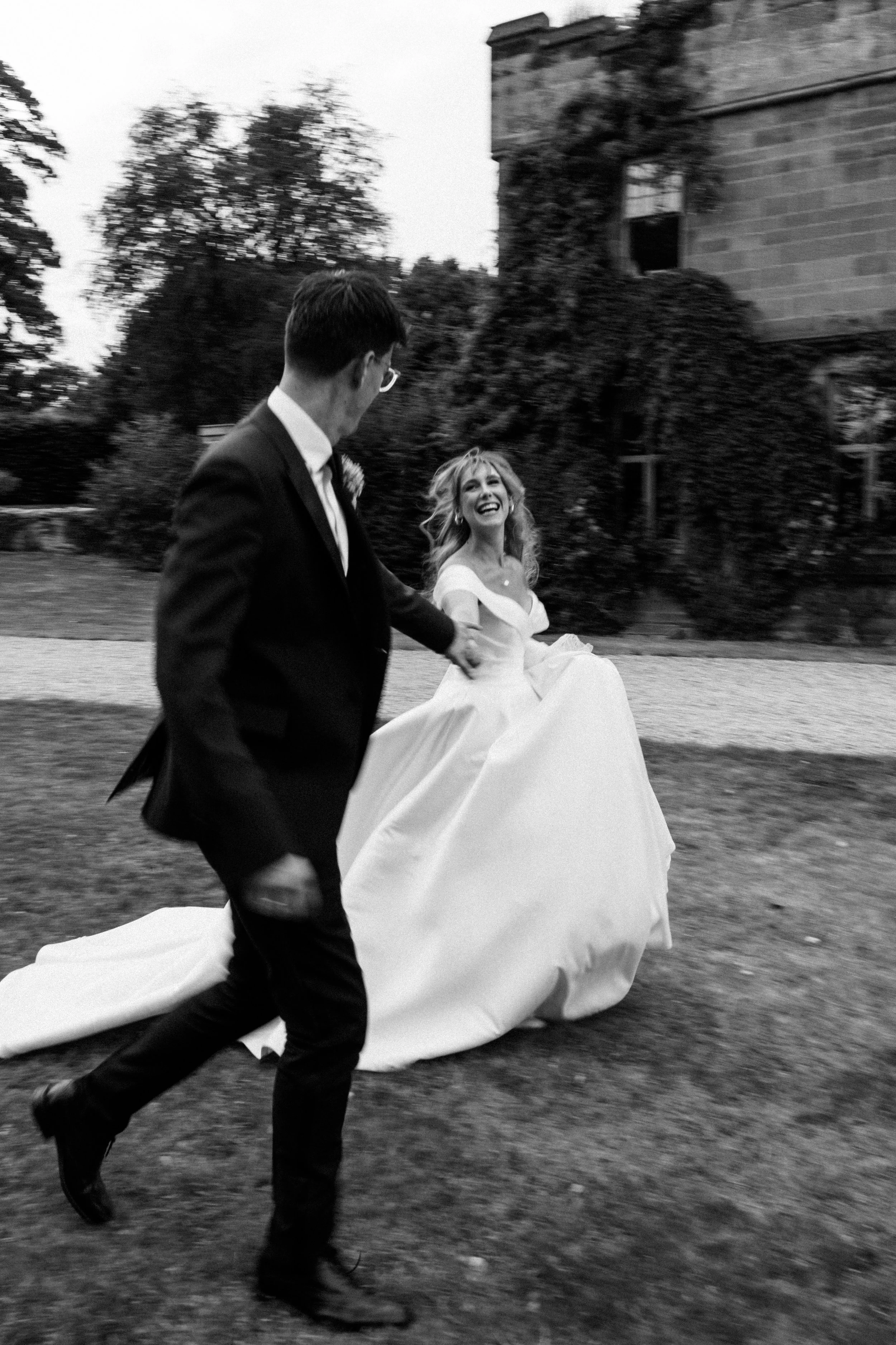 Black and white photo of a couple in wedding attire running outdoors, with the bride smiling and the groom in a suit and glasses, near a building covered in ivy.