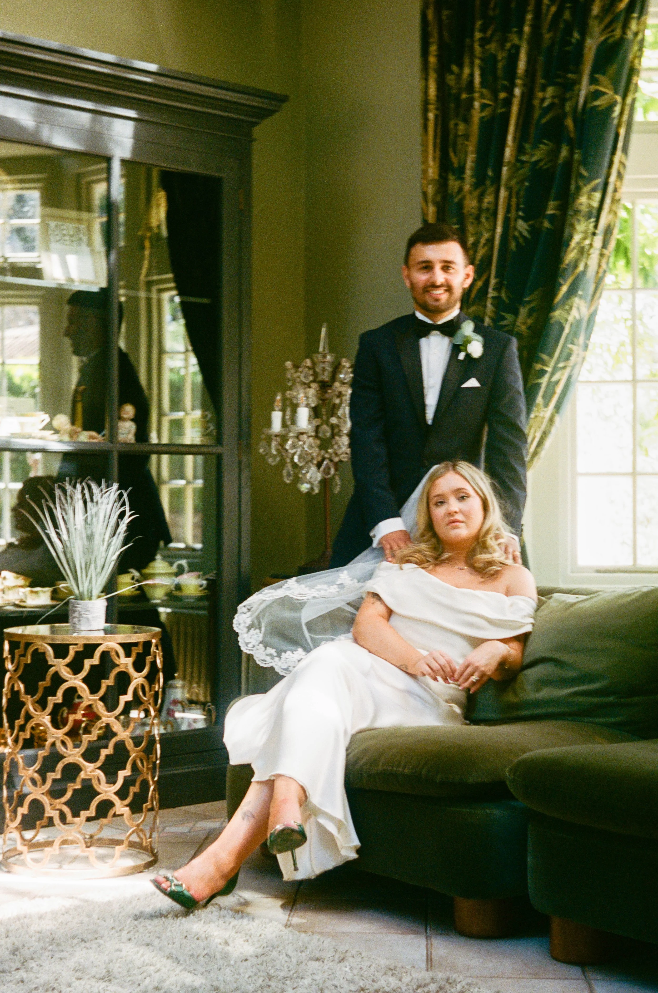 A man in a tuxedo standing behind a woman in a wedding dress who is sitting on a dark green sofa. The woman has blond hair and is wearing high heels. The scene is in a brightly lit room with large windows, curtains, and decorative items.