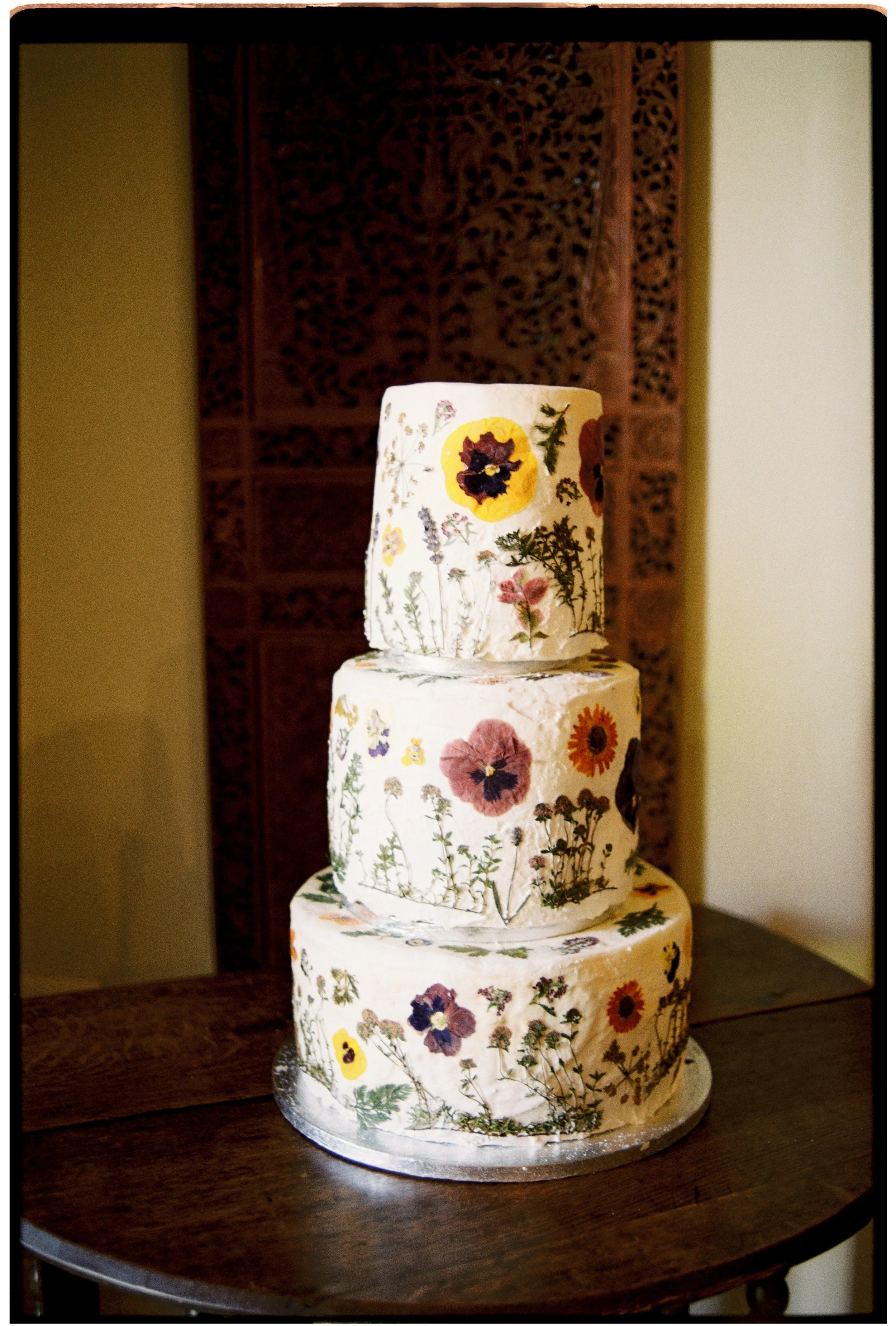 A three-tiered wedding cake decorated with edible flowers and greenery, placed on a wooden table with a decorative wooden panel in the background.