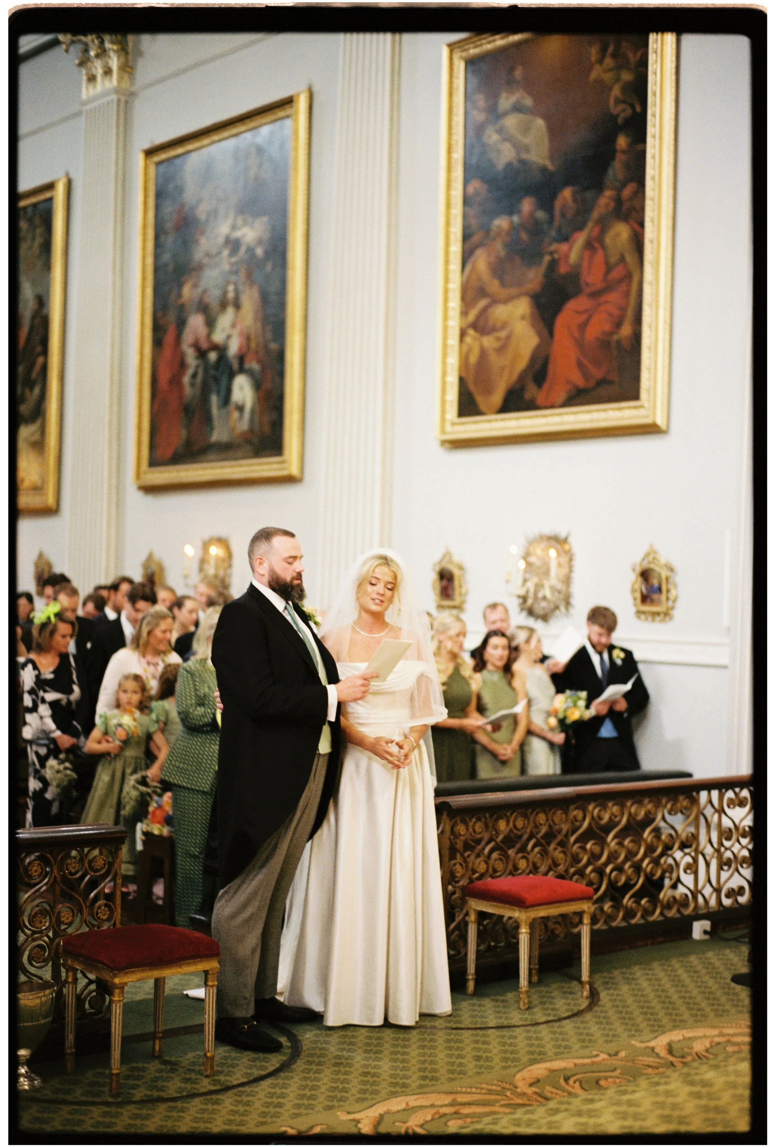 A wedding ceremony inside a church with a bride and groom standing before an officiant. The church is decorated with large framed paintings on the wall and ornate gold accents. Guests are seated in the background.