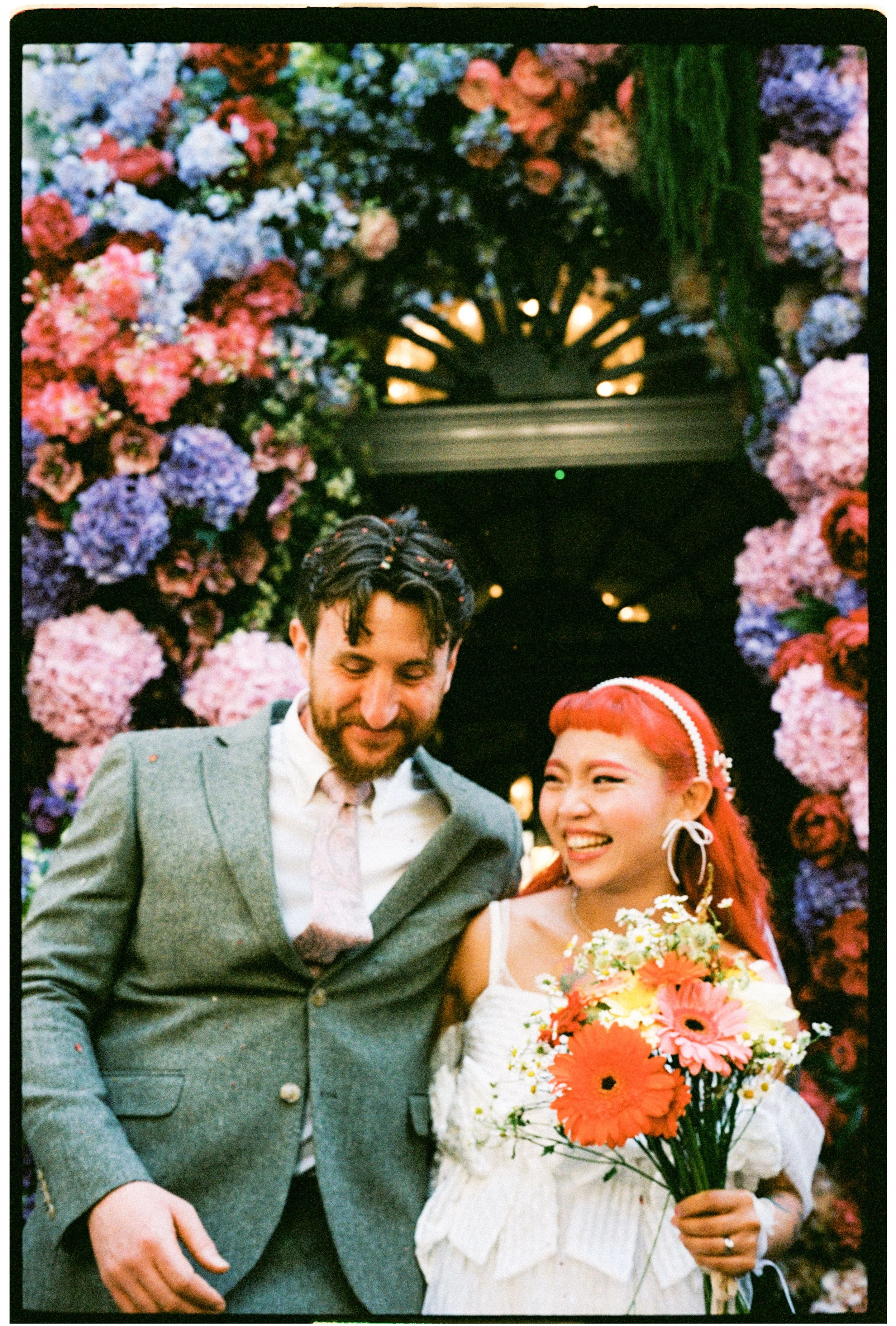 A smiling couple standing side by side during a wedding celebration, surrounded by colorful pink, purple, and blue hydrangea flowers.