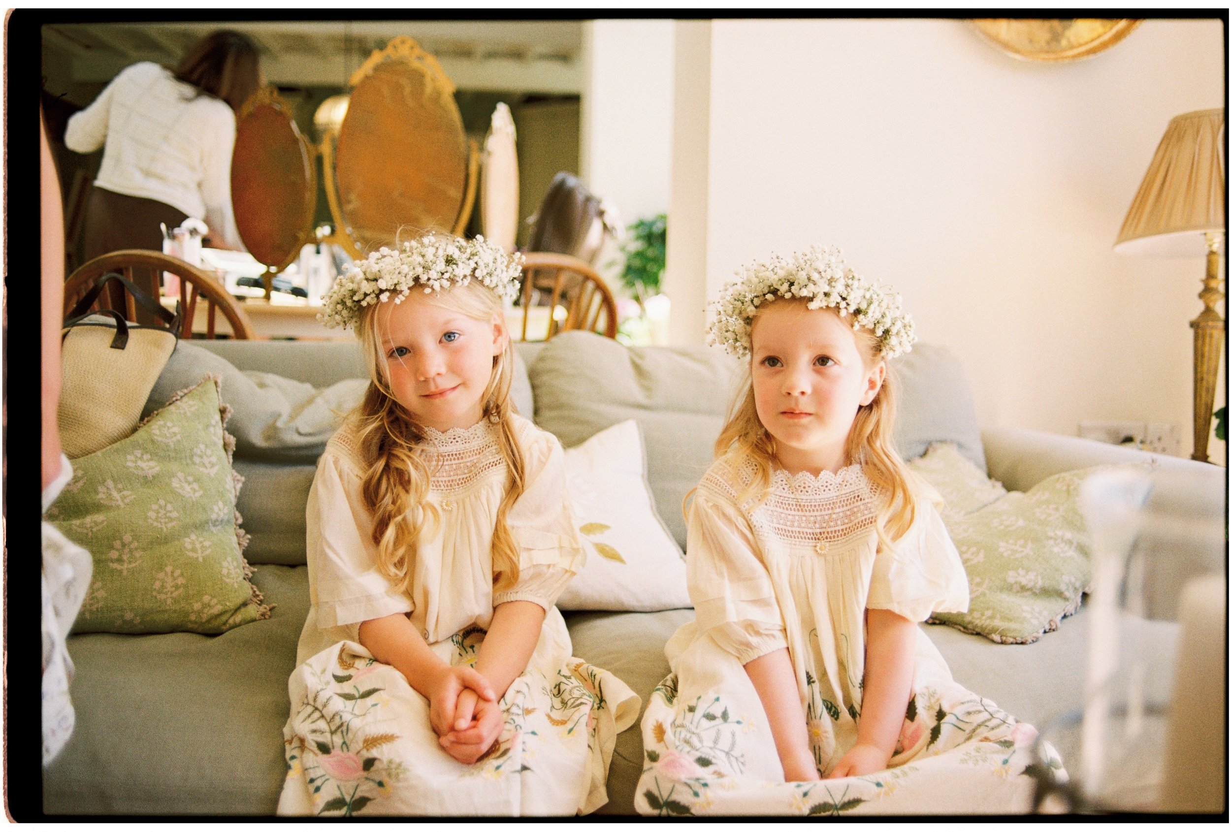 Two young girls sitting on a couch with floral crowns on their heads in a cozy living room.
