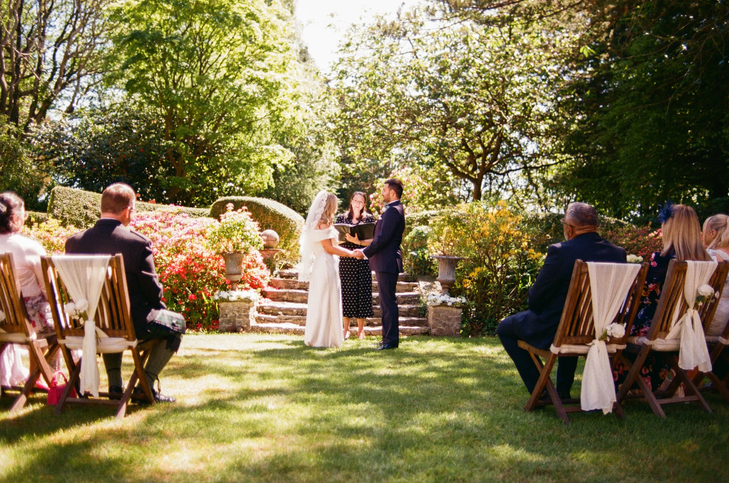 A wedding ceremony outdoors in a garden, with a couple holding hands and exchanging vows in front of an officiant, surrounded by guests seated on wooden chairs amid trees and flowers.