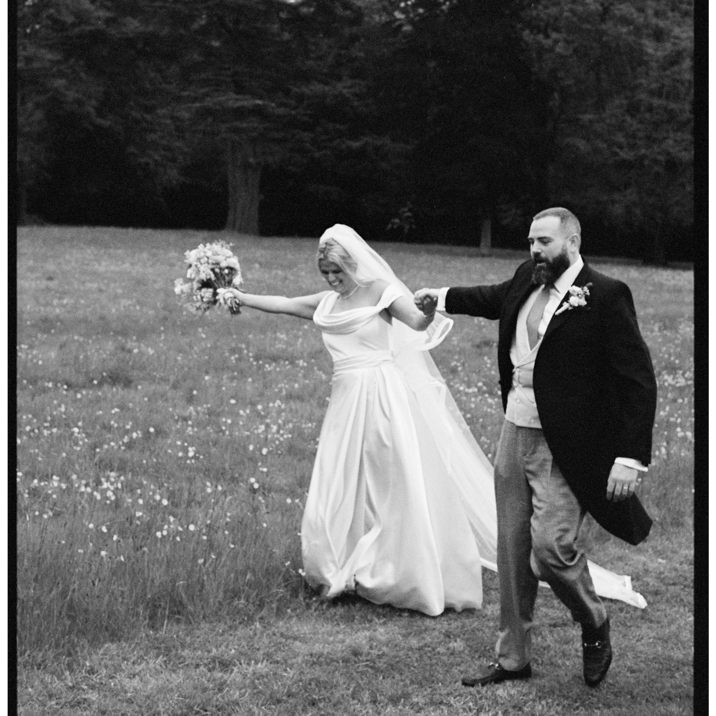 A bride and groom holding hands and running on grass during their wedding, with the bride smiling and holding a bouquet, and the groom dressed in a tuxedo with a beard.
